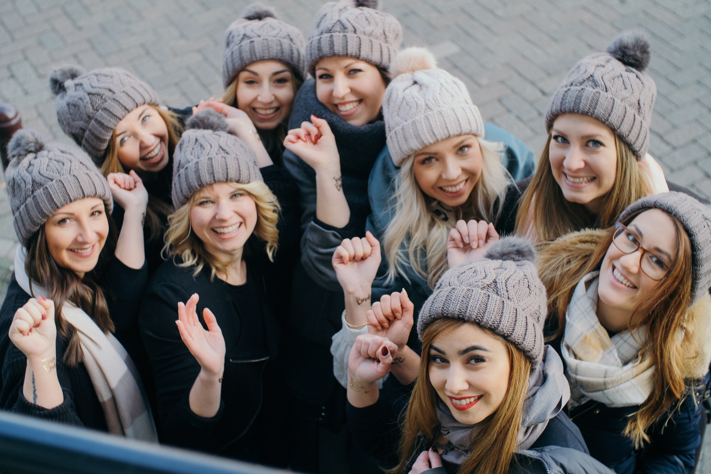Friends at the hen party looking up and raising their fists, gray pompom beanies and scarves, seen from above on the cobbles