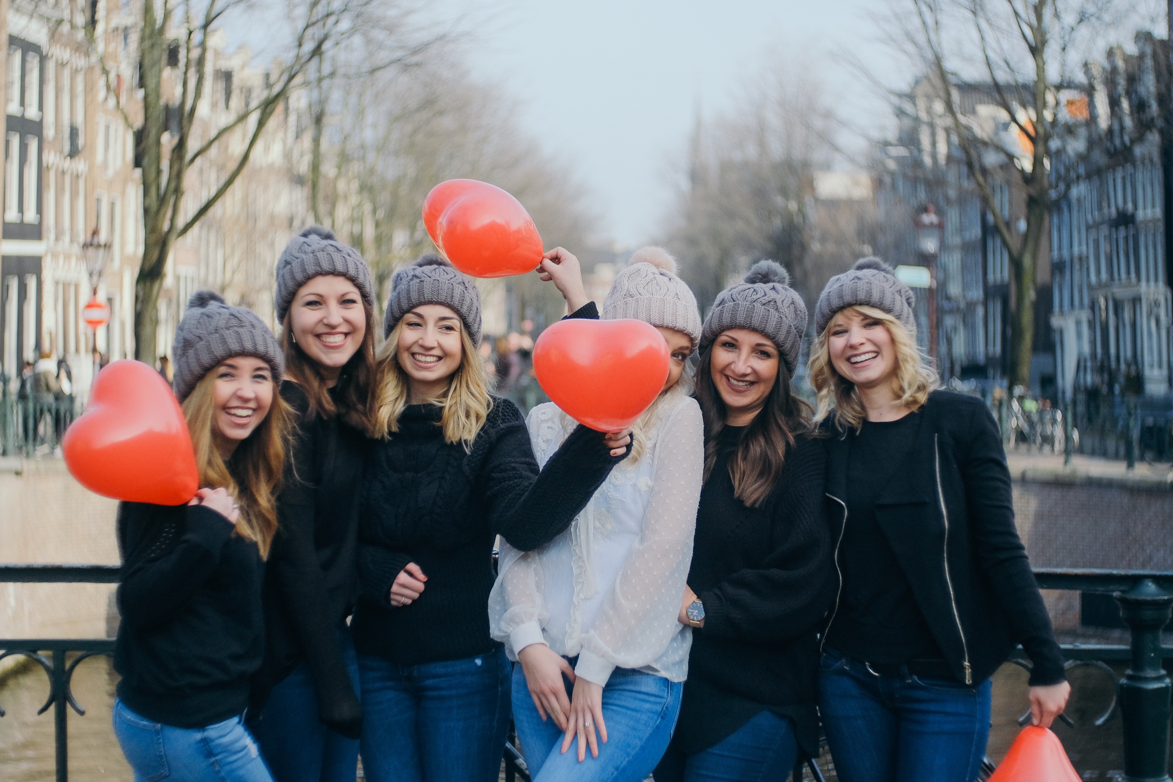 Friends in gray knit beanies holding red heart balloons on a canal bridge, one balloon in front of the bride's face at their winter hen party