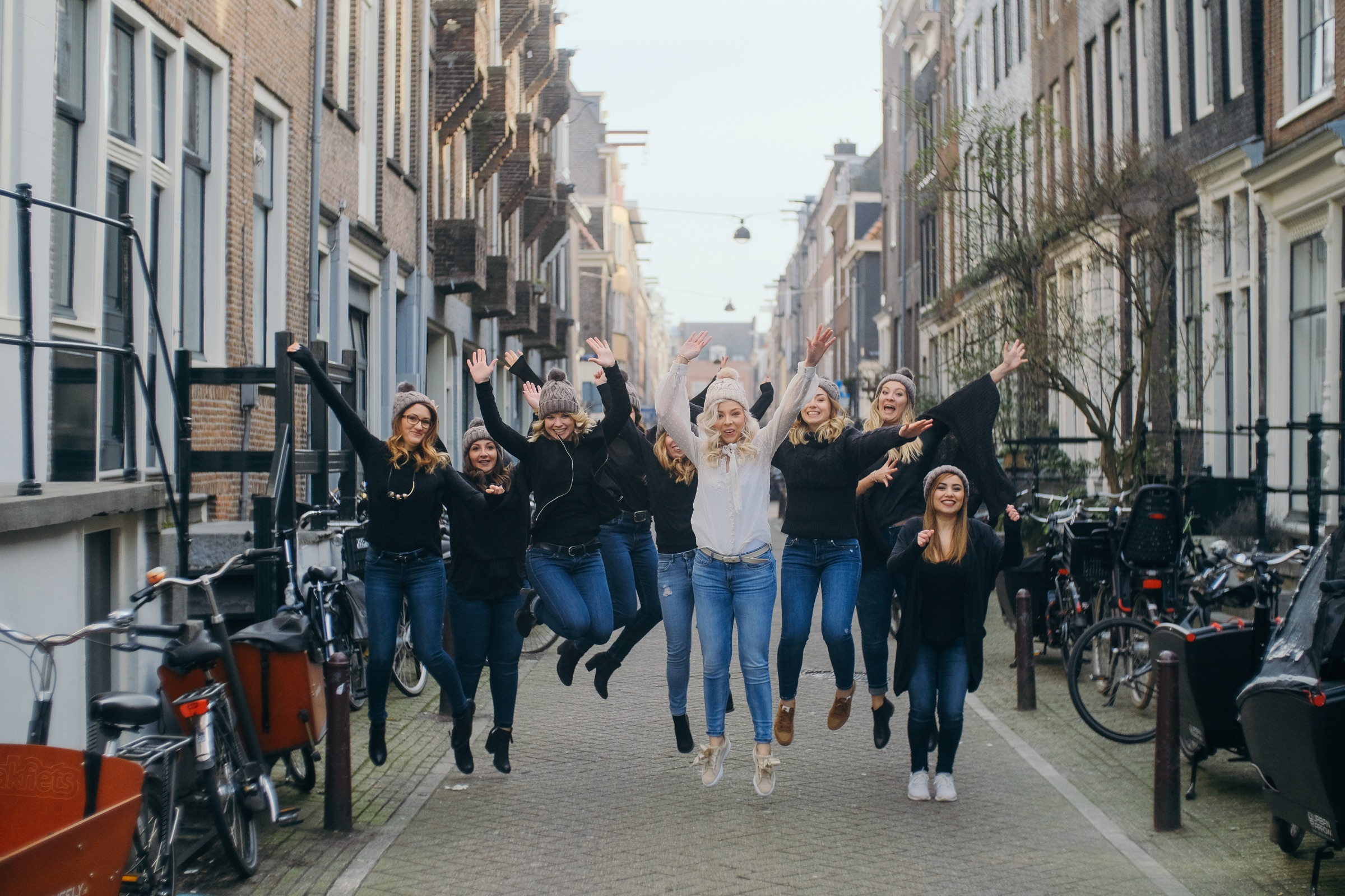 Hen party friends jumping with their arms up in a narrow street lined with bikes, the bride in white in the middle