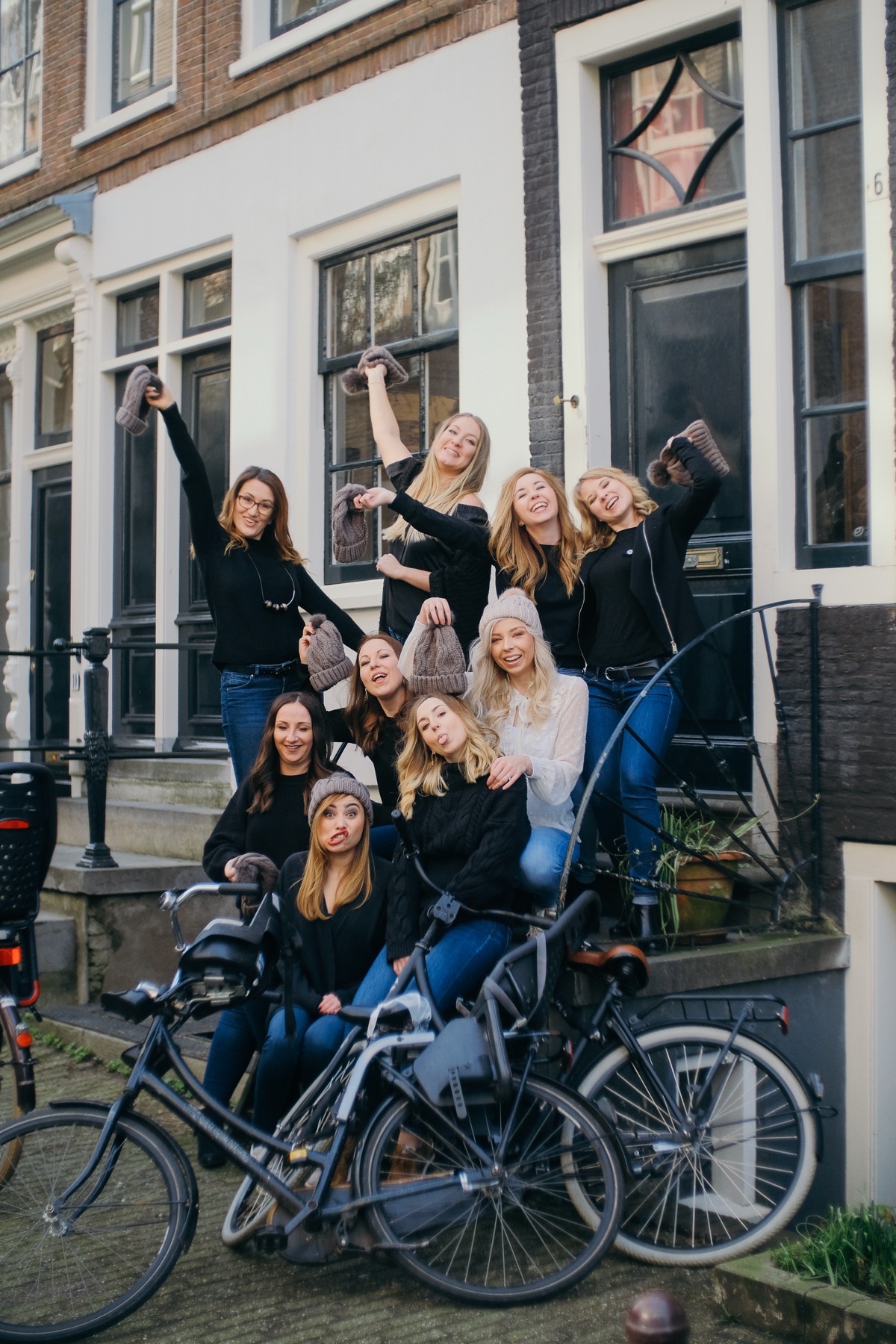 Hen party group crowded onto the stoop of a white canal house, waving gray beanies in the air above parked bikes