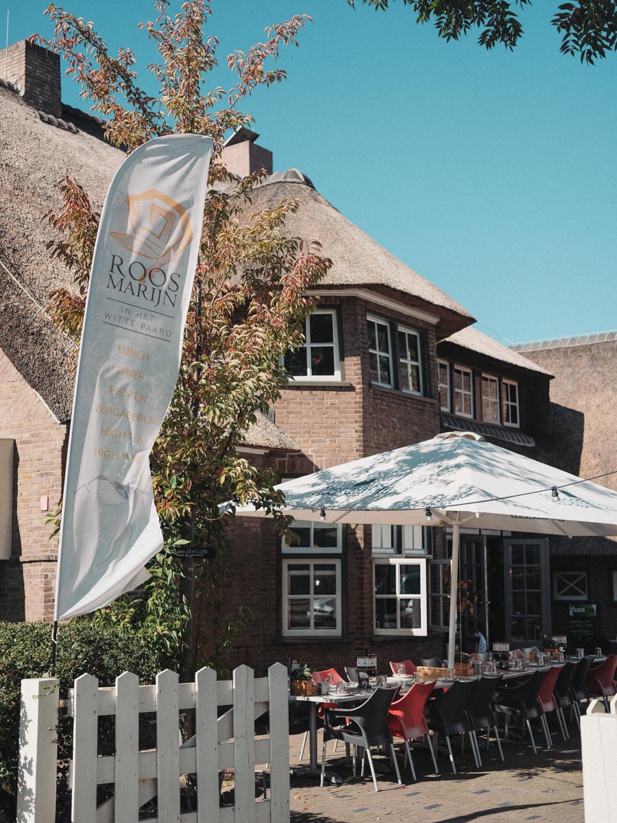Thatched brick restaurant with a white picket fence and parasols on its terrace, the restaurant’s tall white flag by the door