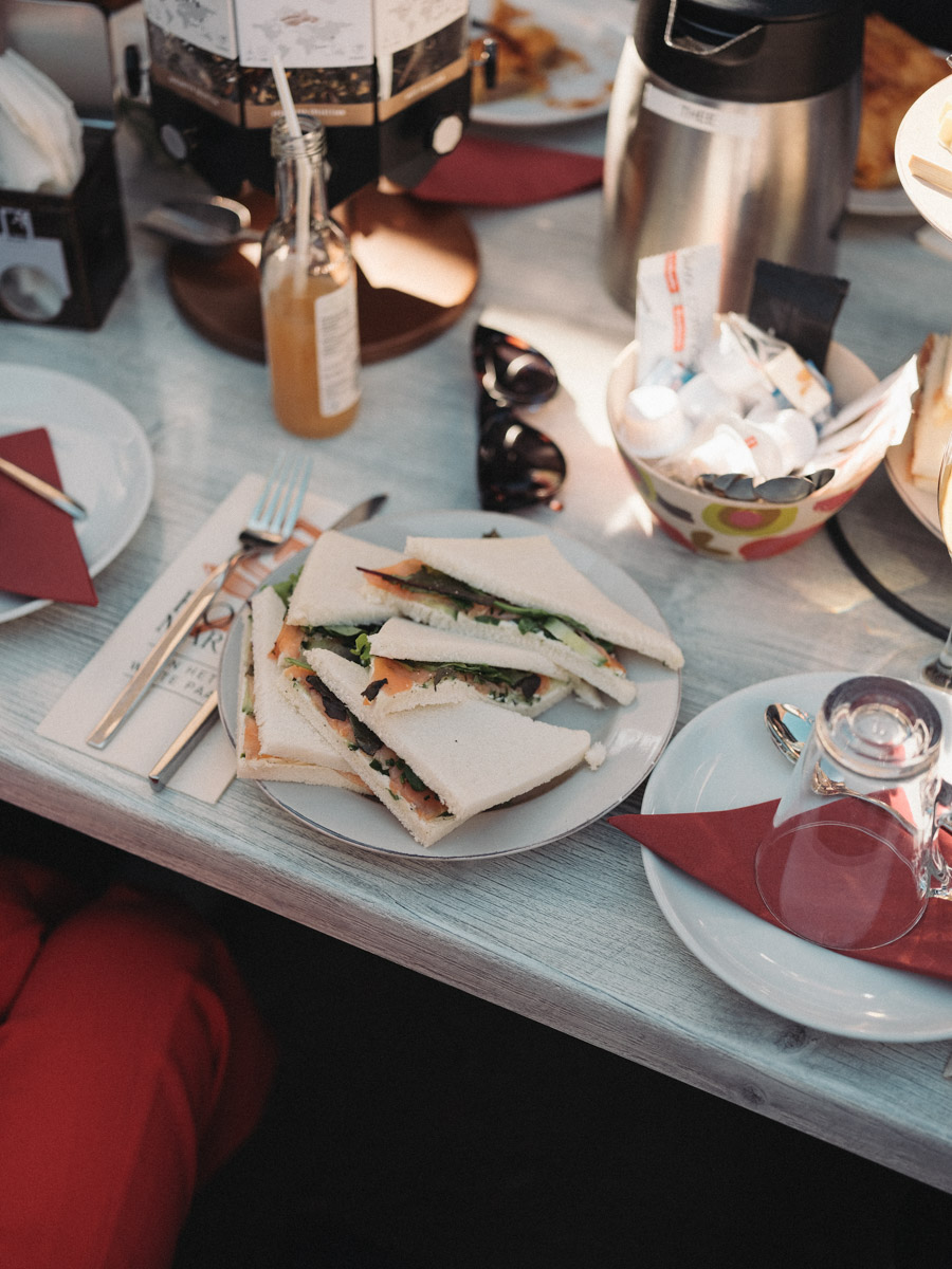 Plate of sandwiches cut into triangles on a white table in the sun, a bottle of juice and a bowl of sugar sachets beside it