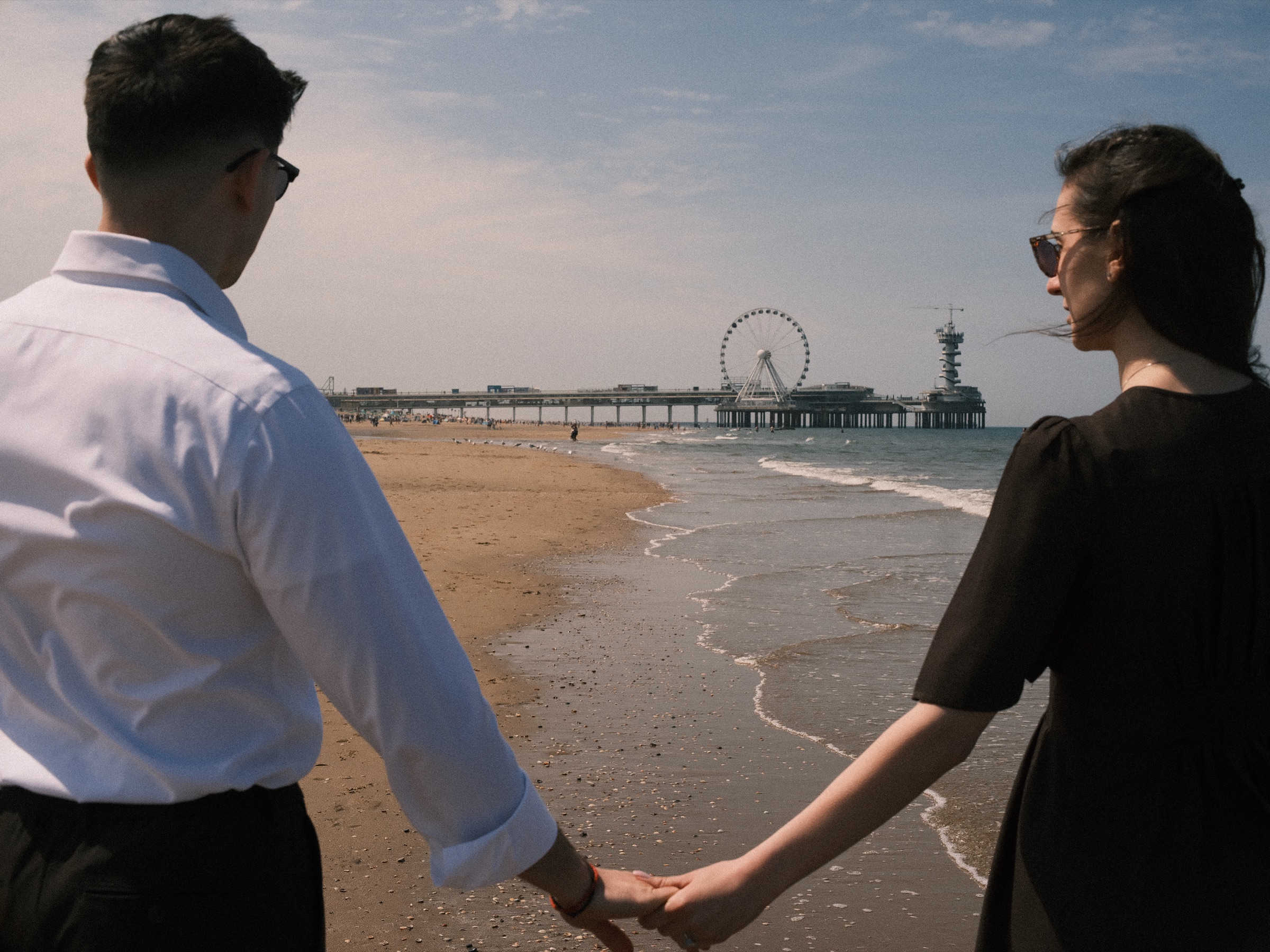 Seen past his shoulder, the couple holds hands at the water with the Pier and its Ferris wheel between them on Scheveningen beach