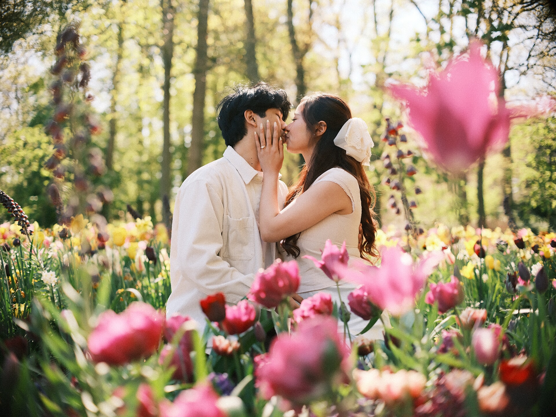 Couple kissing among pink tulips at Keukenhof, her hand on his cheek and yellow daffodils glowing in the woods behind
