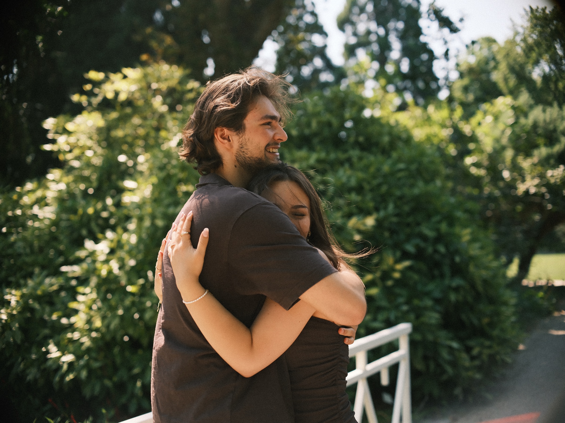 Long tight hug on the white bridge, his face turned toward the lens with a wide smile, green shrubs all around them