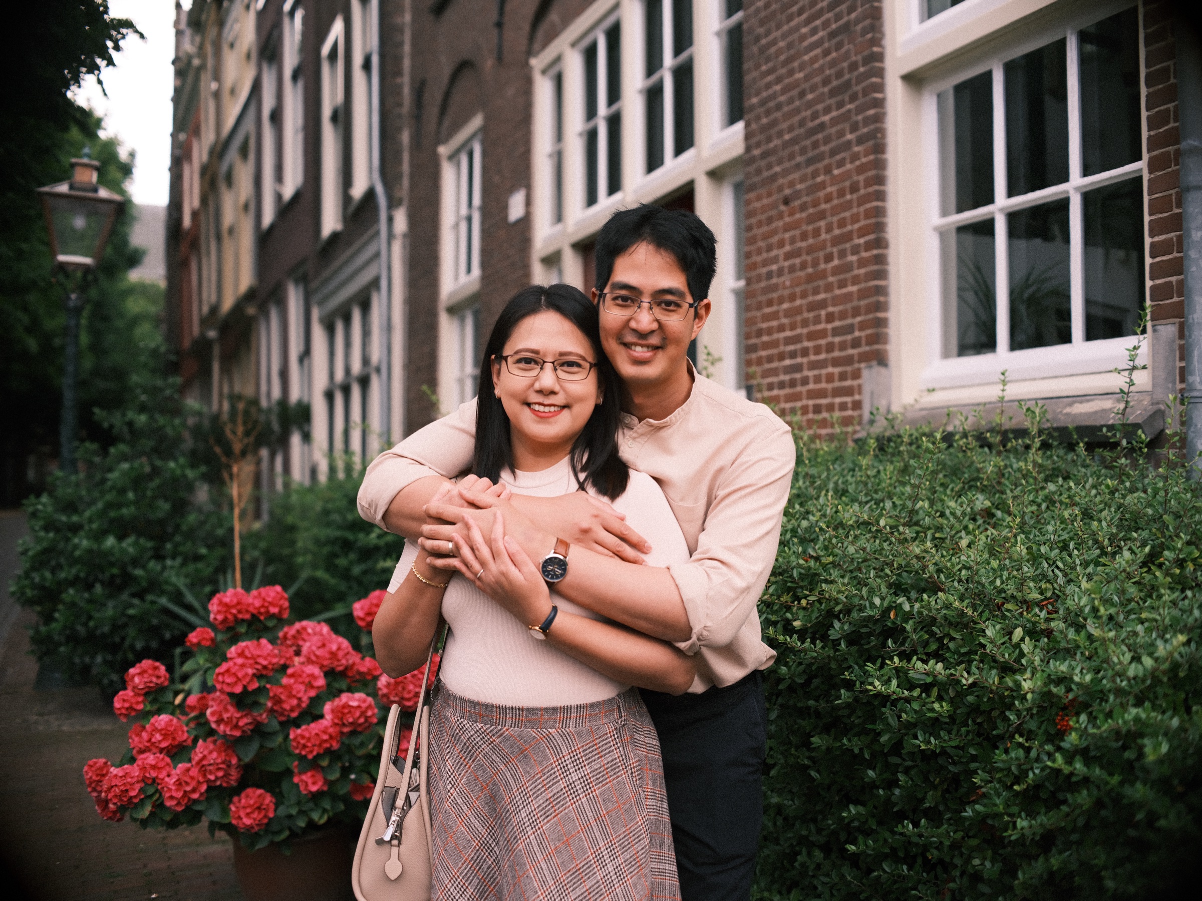 He hugs her from behind and they both smile, red geraniums in a pot and a trimmed hedge beside a tall brick house in Leiden