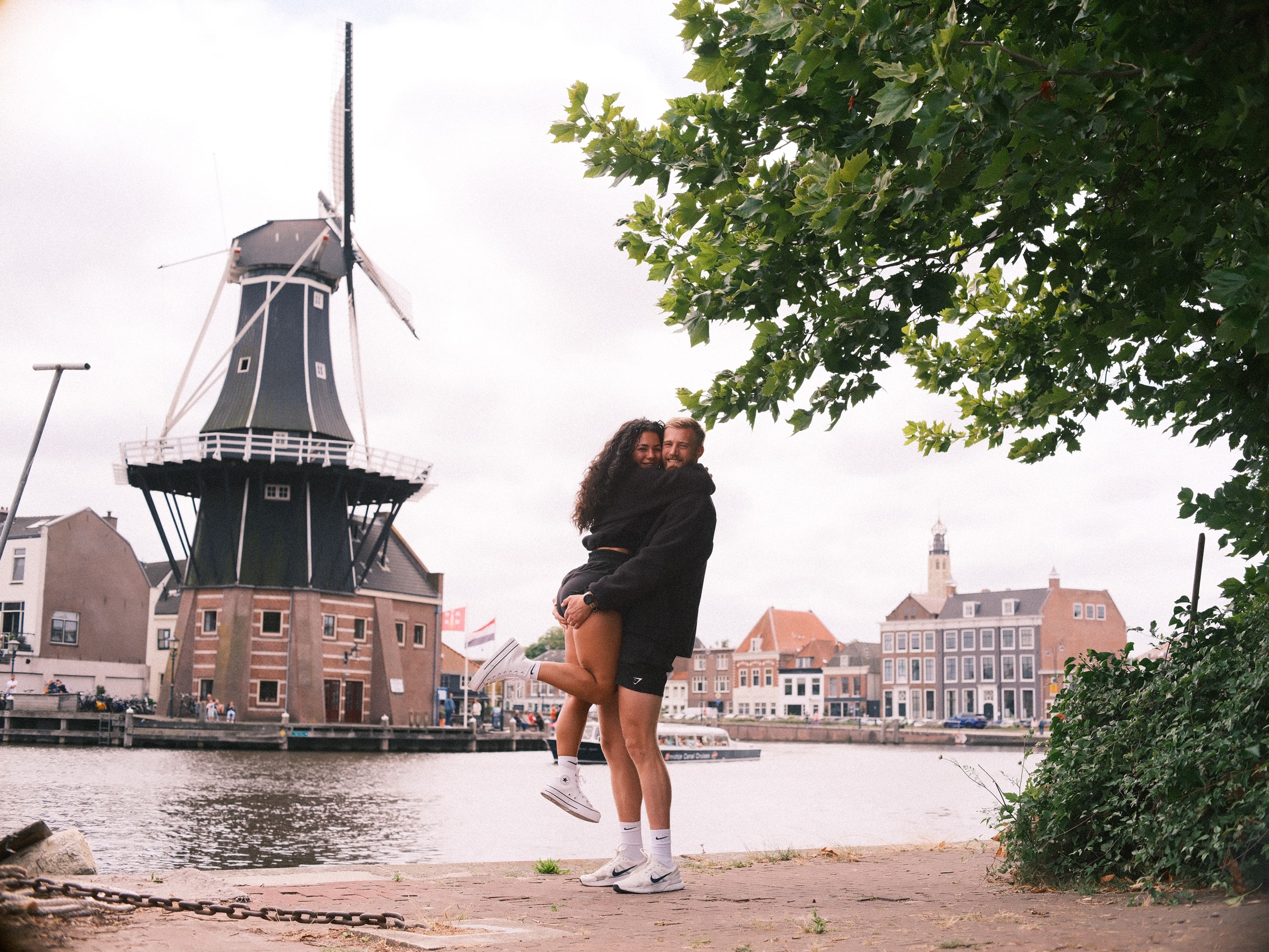 Zij springt hem in de armen op de kade tegenover molen De Adriaan, de molen omlijst door een boom en een bleke zomerlucht
