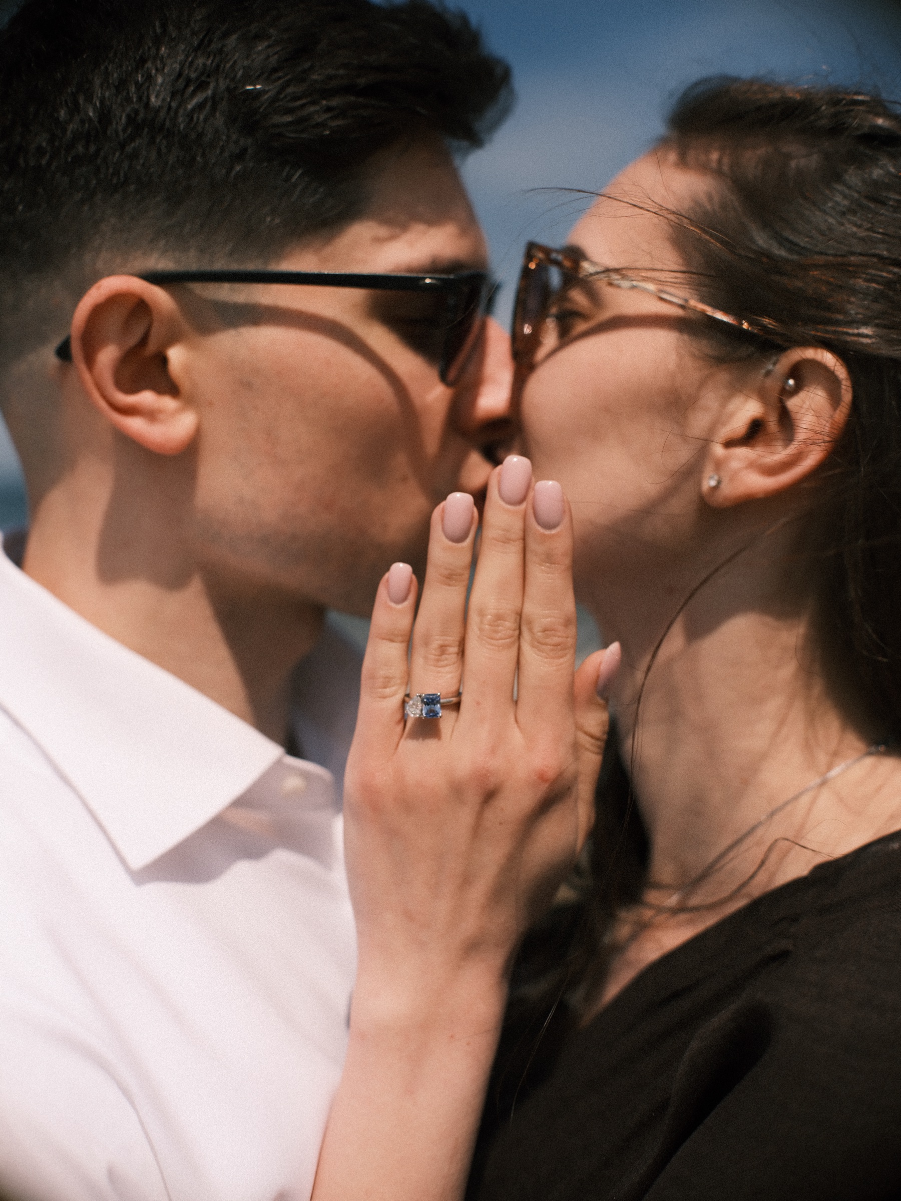 Close kiss behind her raised hand, the blue engagement ring on her finger and both of them in sunglasses