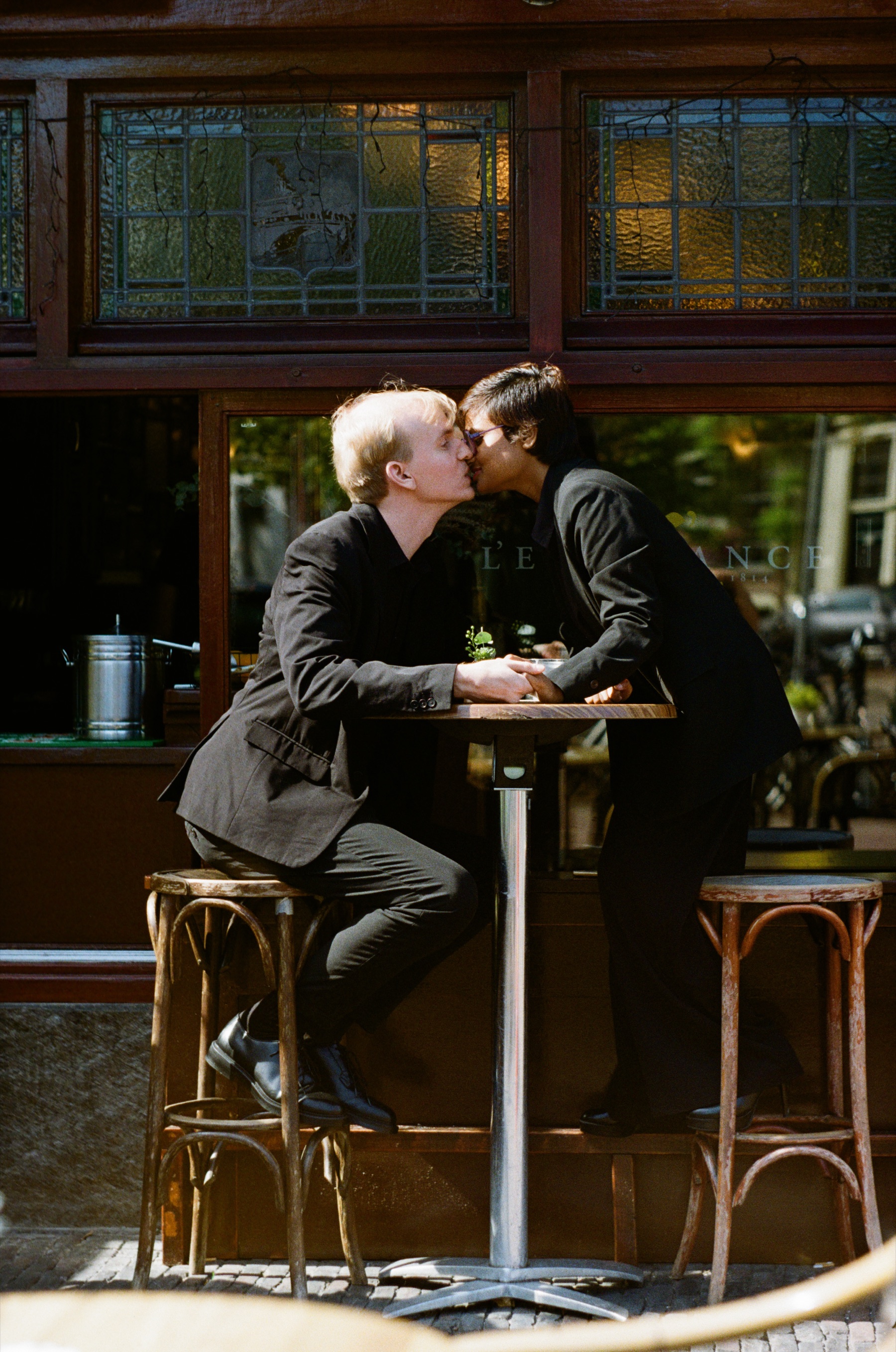 Kiss over a café table on tall stools, the warm reds and browns of the wooden storefront glowing on Kodak Gold in Leiden