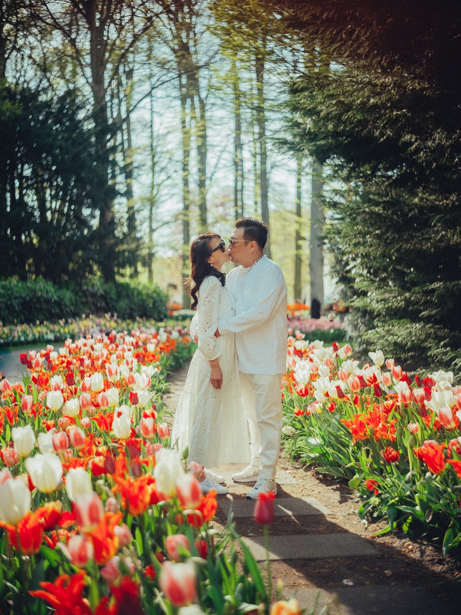 Man in het wit kust de vrouw op de wang terwijl ze tussen rode en witte tulpen lopen, met zonlicht door de bomen