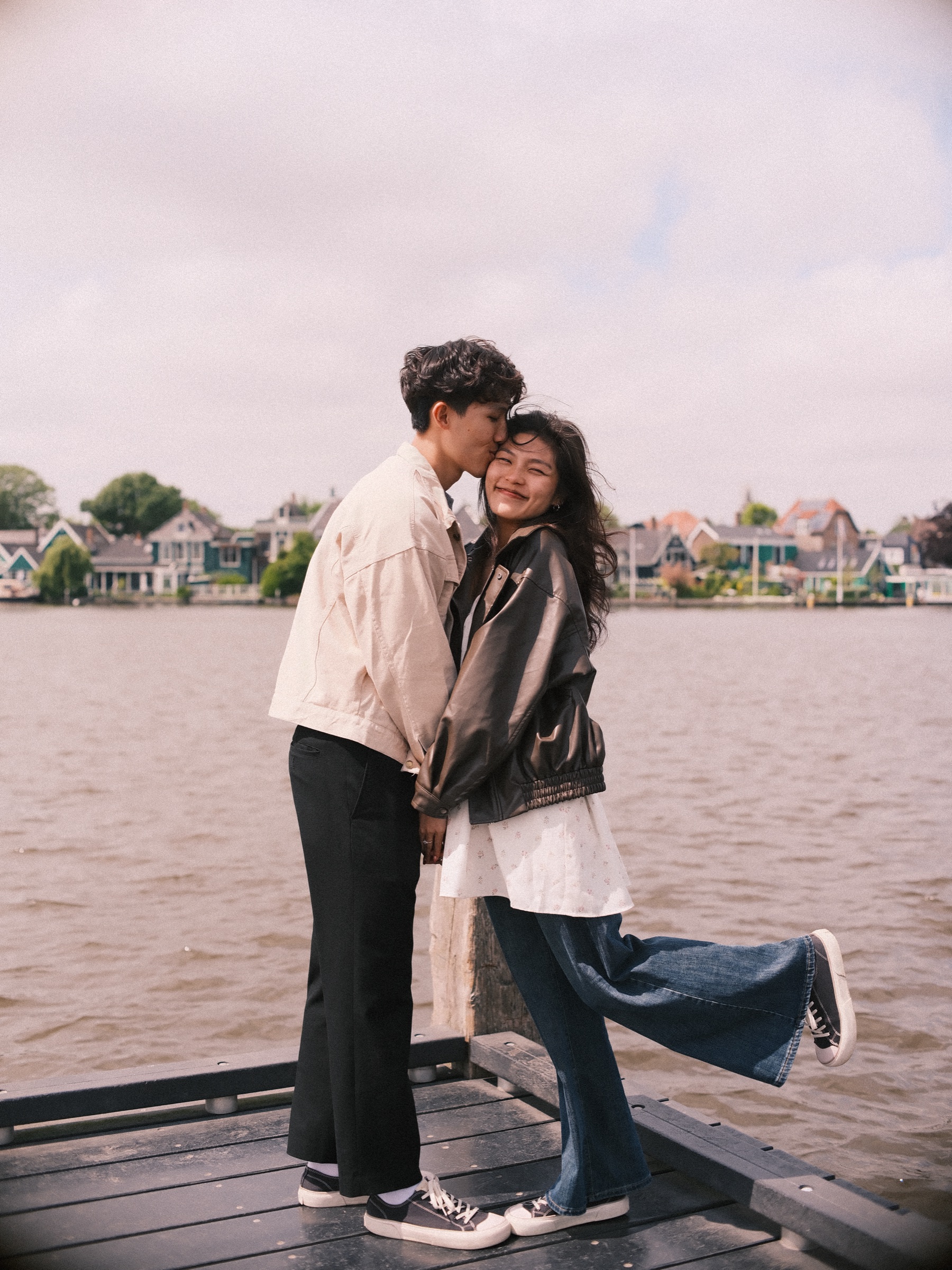 Couple kissing at the end of a jetty while she kicks one sneaker up behind her, the river and green houses on the far bank behind
