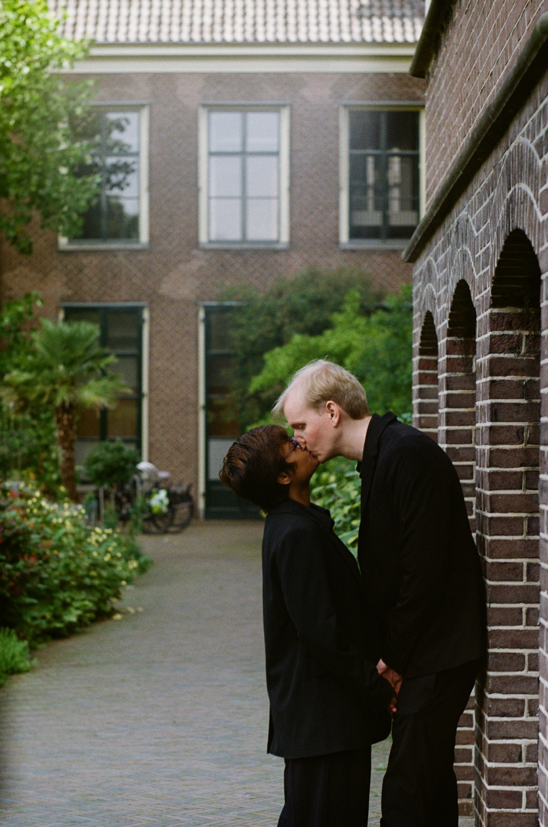 Kiss in a quiet green courtyard in Leiden, plants and a small palm behind them, a soft frame from a roll of Kodak Gold