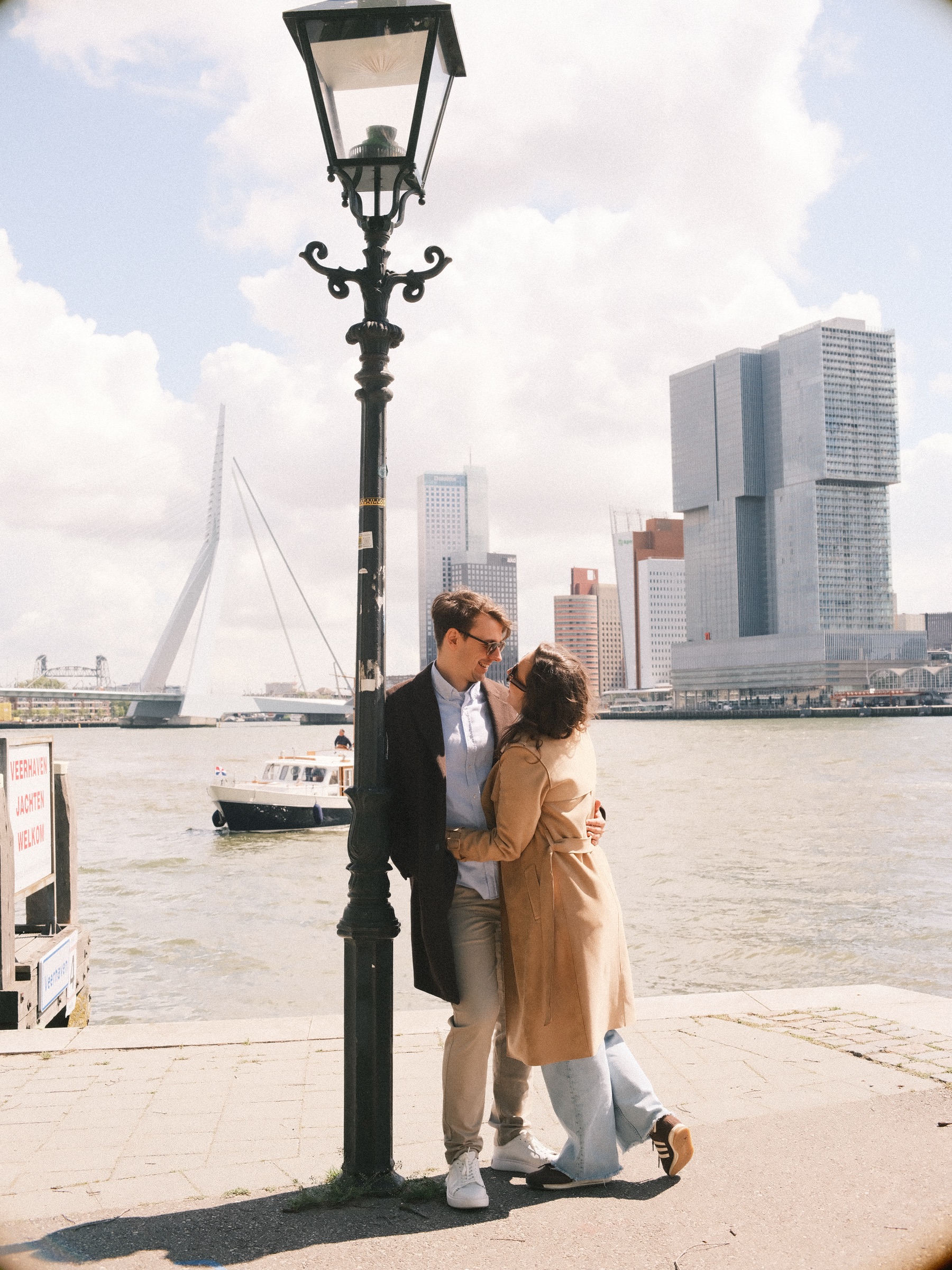 Kiss beside an old black lamppost where the Veerhaven meets the Maas, a water taxi passing and the Erasmusbrug under white clouds