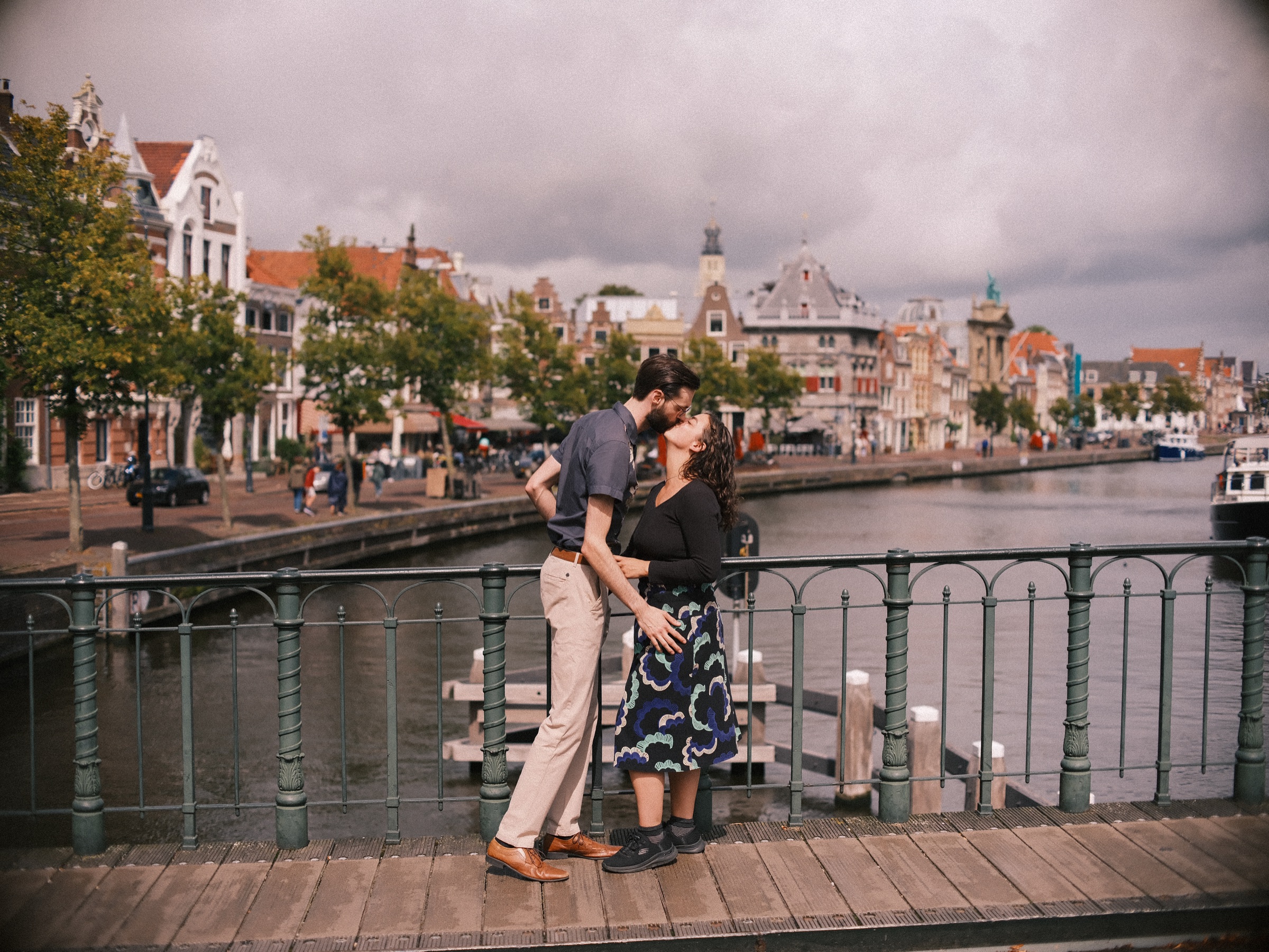 Stel kust op een brug met een groen ijzeren hek boven het Spaarne, met oude gevels en boten onder een zware grijze lucht