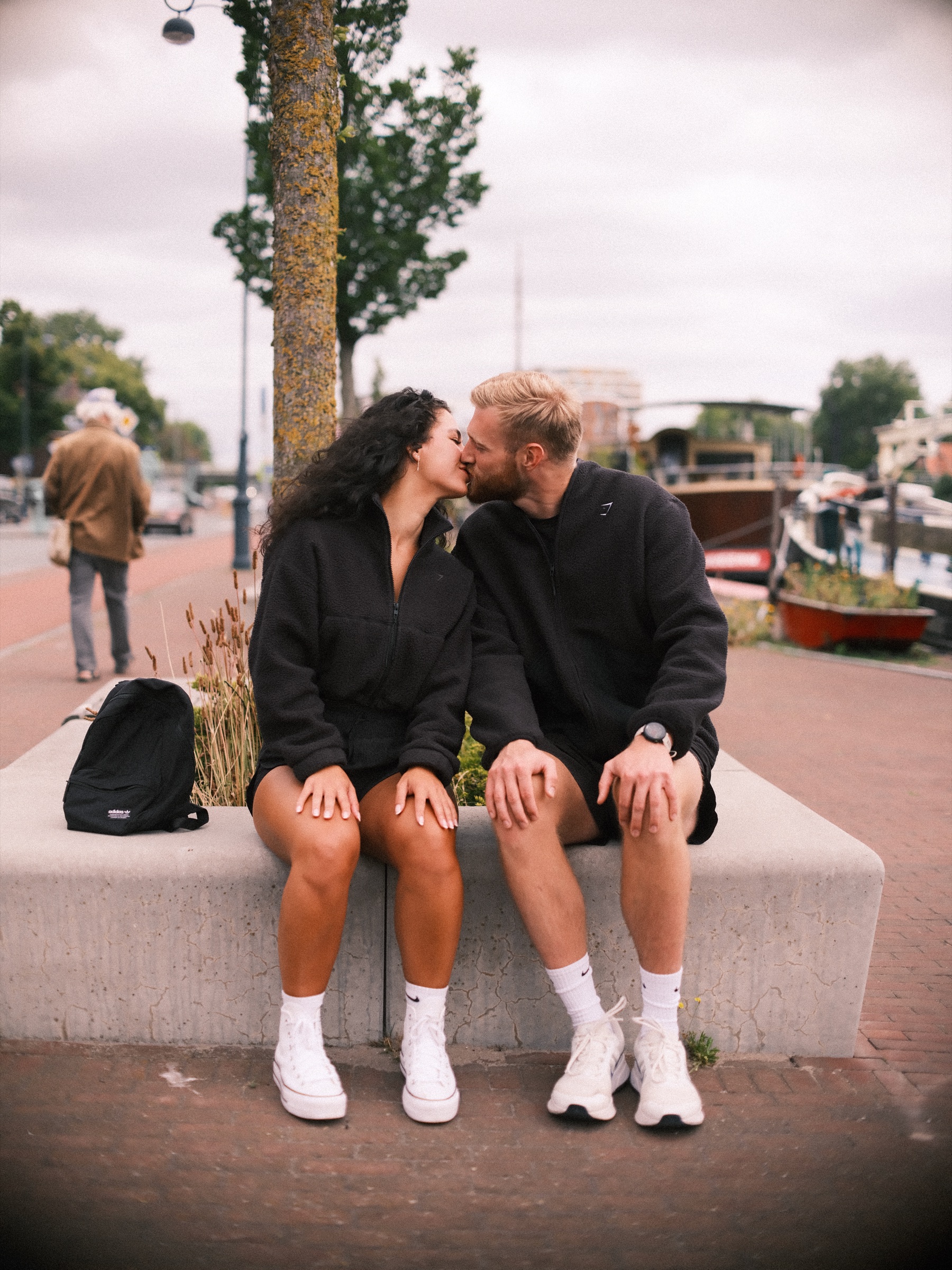 Vrouw geeft haar vriend een kus op de wang op een betonnen bank aan het Spaarne, met een zwarte rugzak naast hen