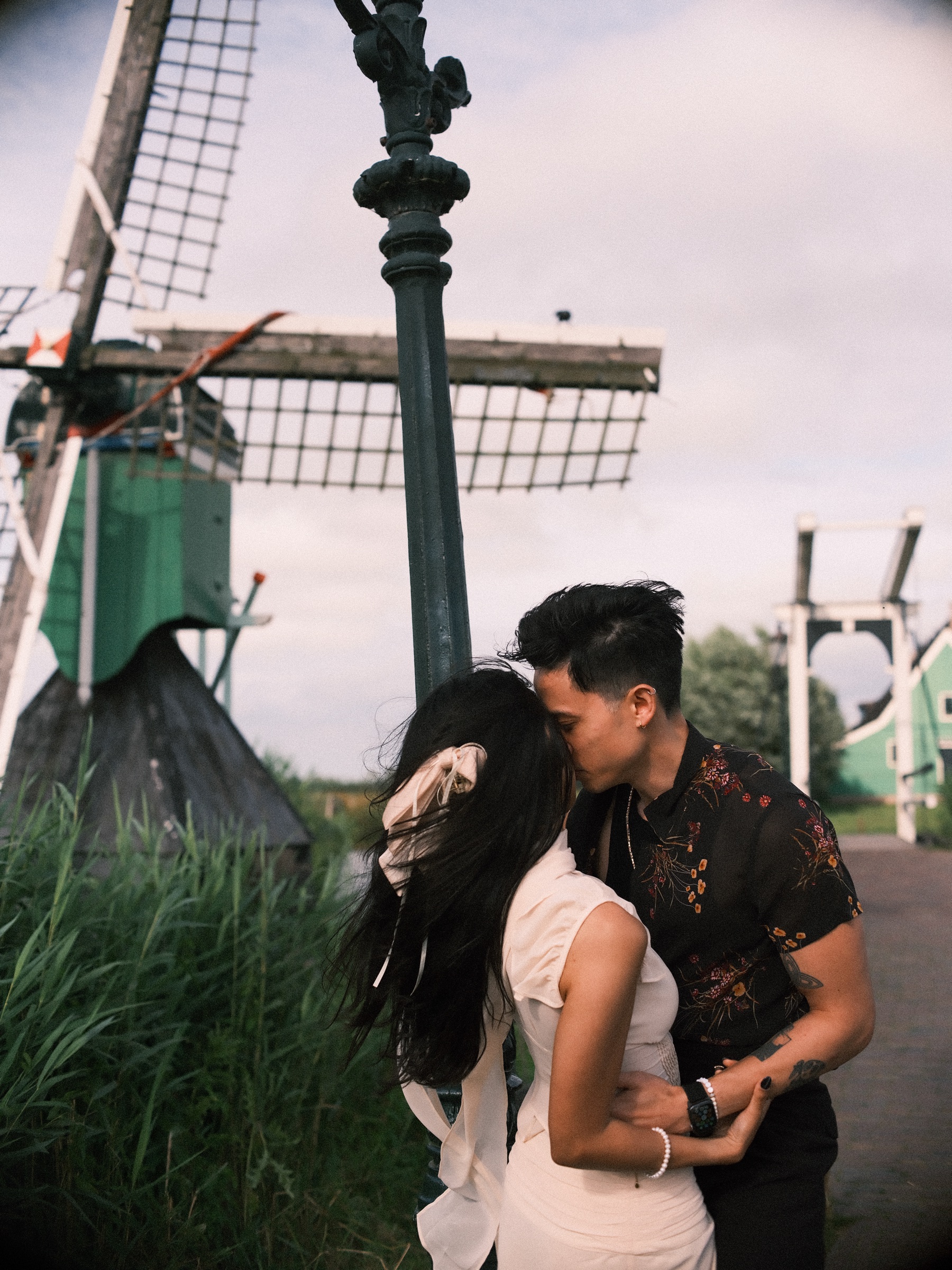 Man kissing her cheek by a lamppost and the sails of a small windmill, her long ribbon and dark hair blowing in the wind