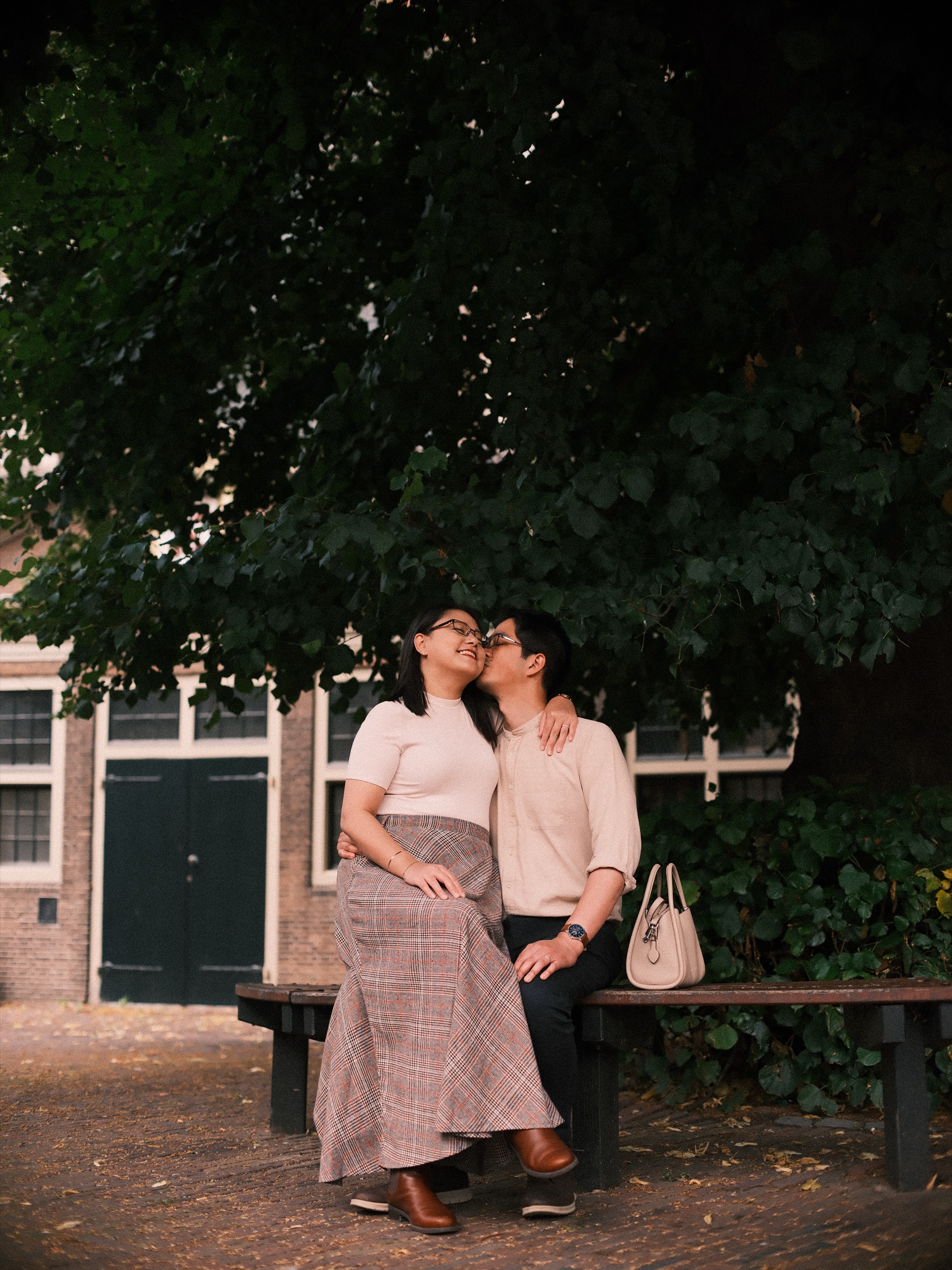 Woman kissing her partner on the cheek on a shady bench, a handbag beside them and dark green doors in the brick behind