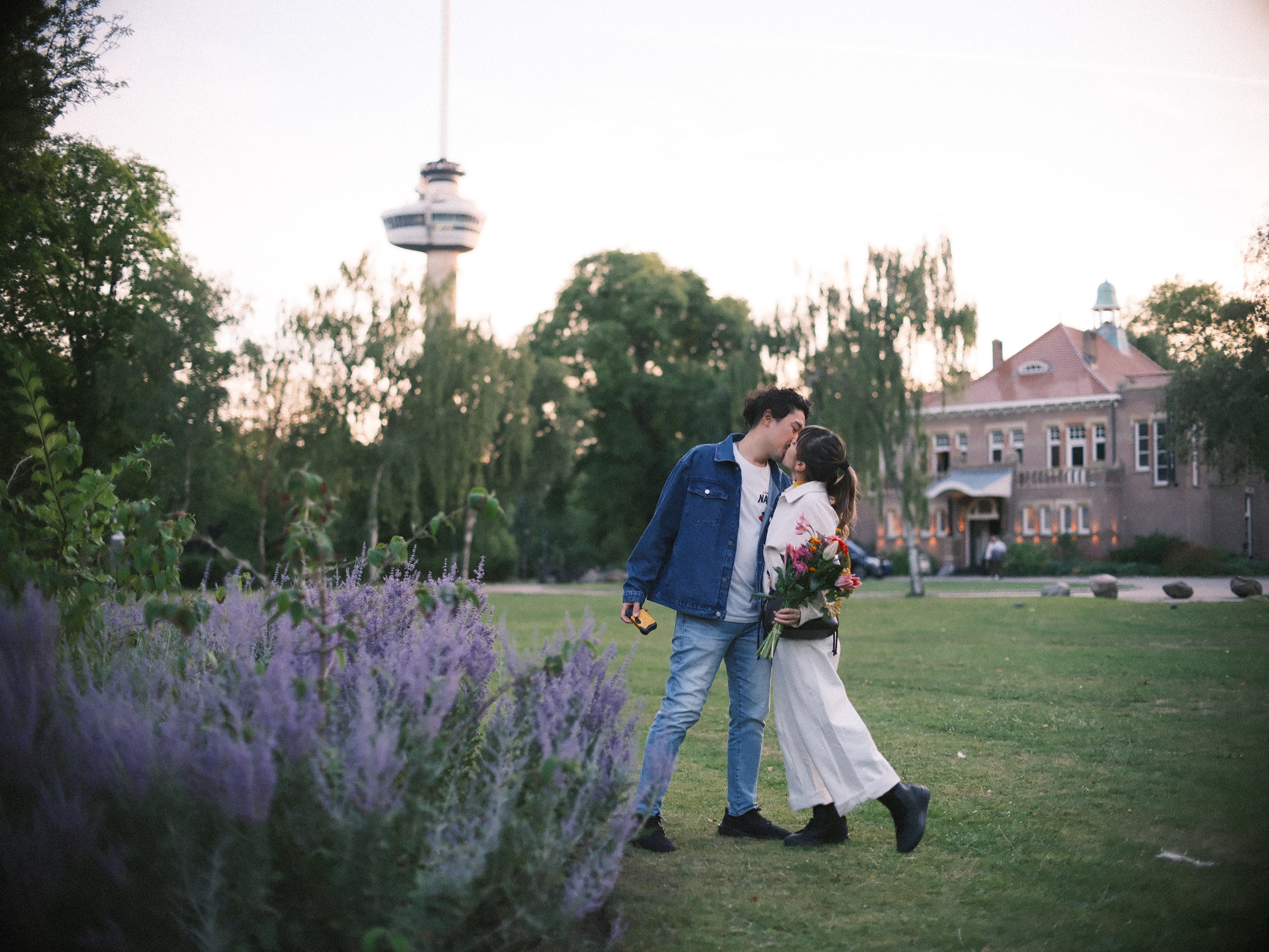 Man geeft haar een kus op de wang terwijl ze over het gras van Het Park lopen, met de Euromast hoog boven de bomen