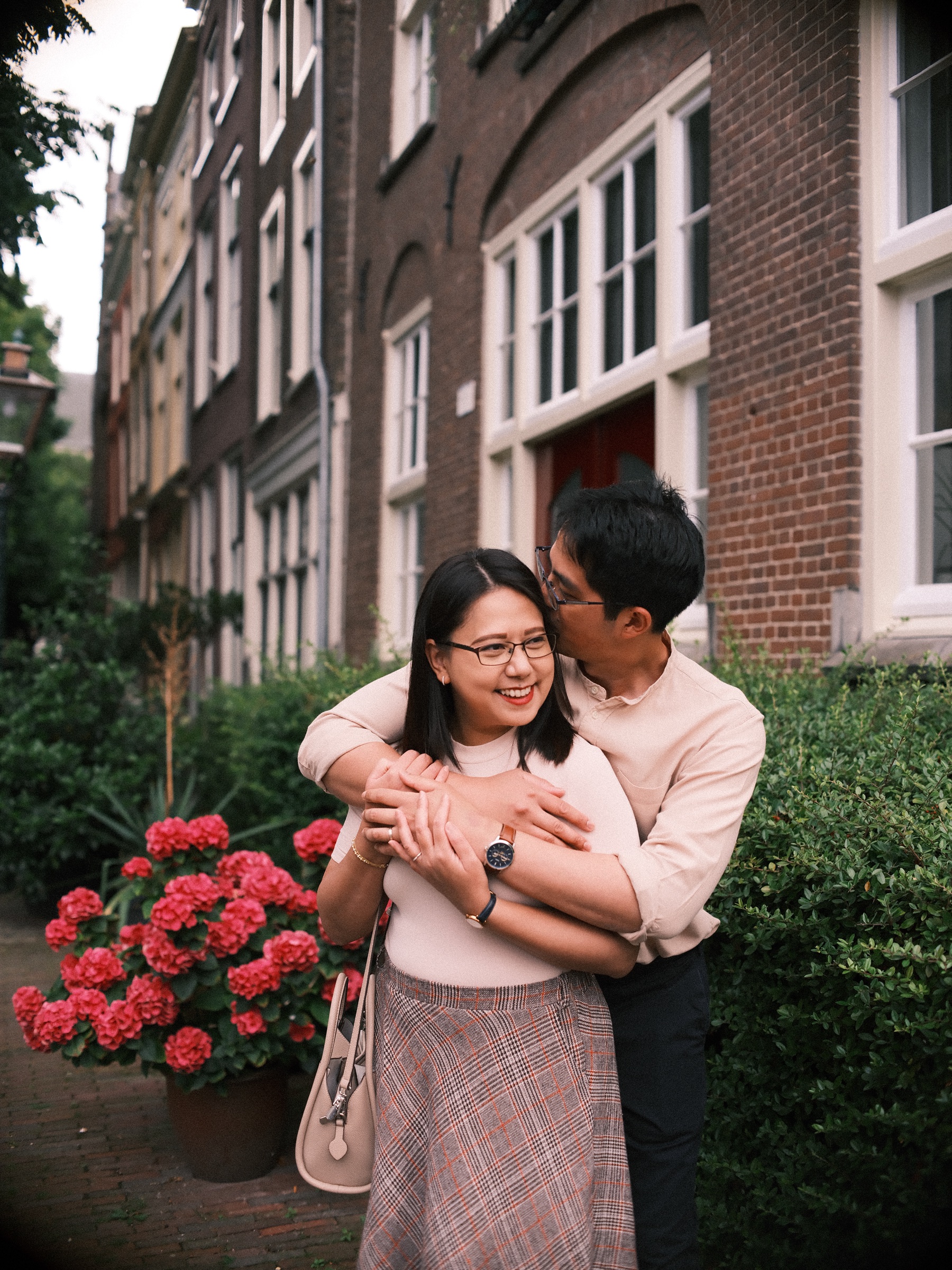 Kiss on the temple in front of a hedge and potted geraniums, her eyes closed in a smile and his watch on the arm around her
