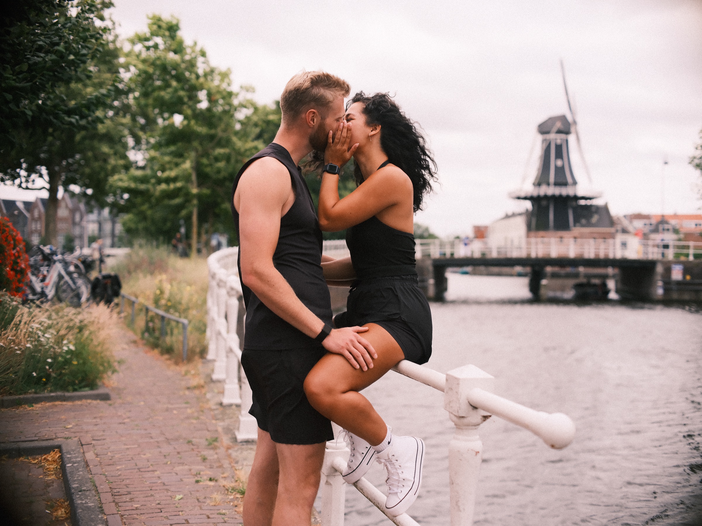 Vrouw zit op een wit hek en kust haar vriend, allebei in zwarte topjes, met de molen en een brug aan de overkant van het water