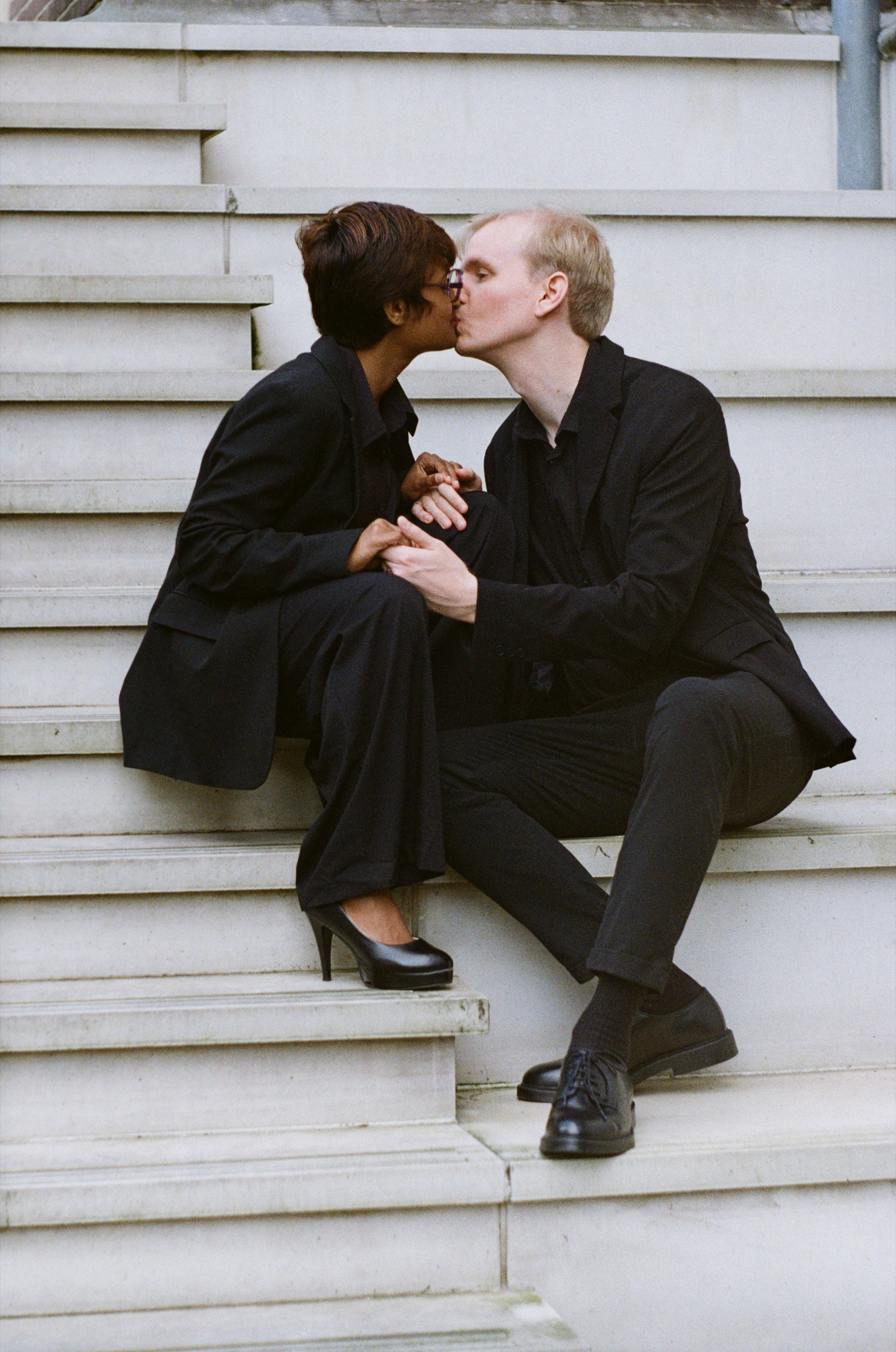 Couple kissing on white stone steps, her black heels and his polished shoes in the sun, the steps bright against their clothes
