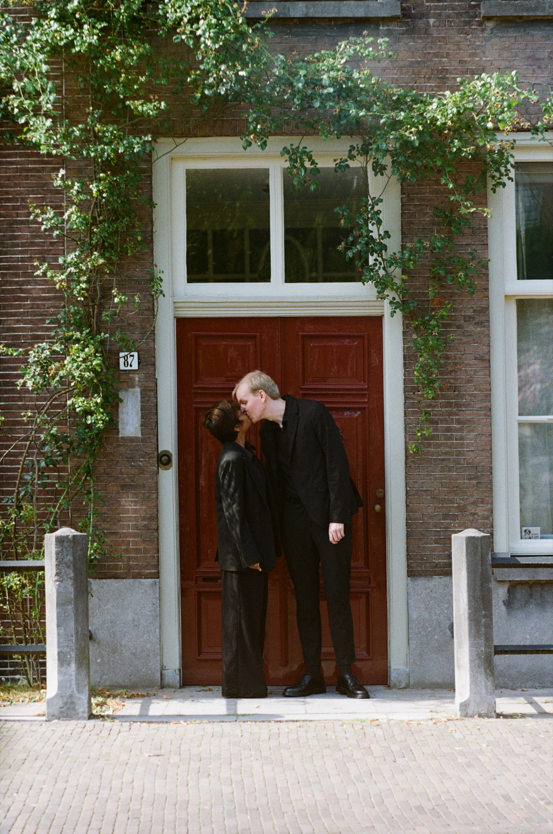 Kiss at a red door under ivy in Leiden, a quiet doorstep frame on Kodak Gold with stone posts on either side of the step