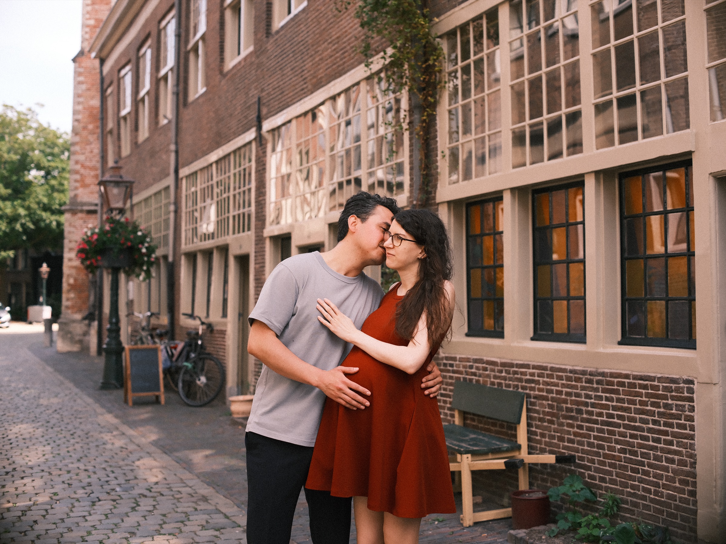 Man kissing the temple of his pregnant partner beside a long brick building with tall windows glowing warm from inside