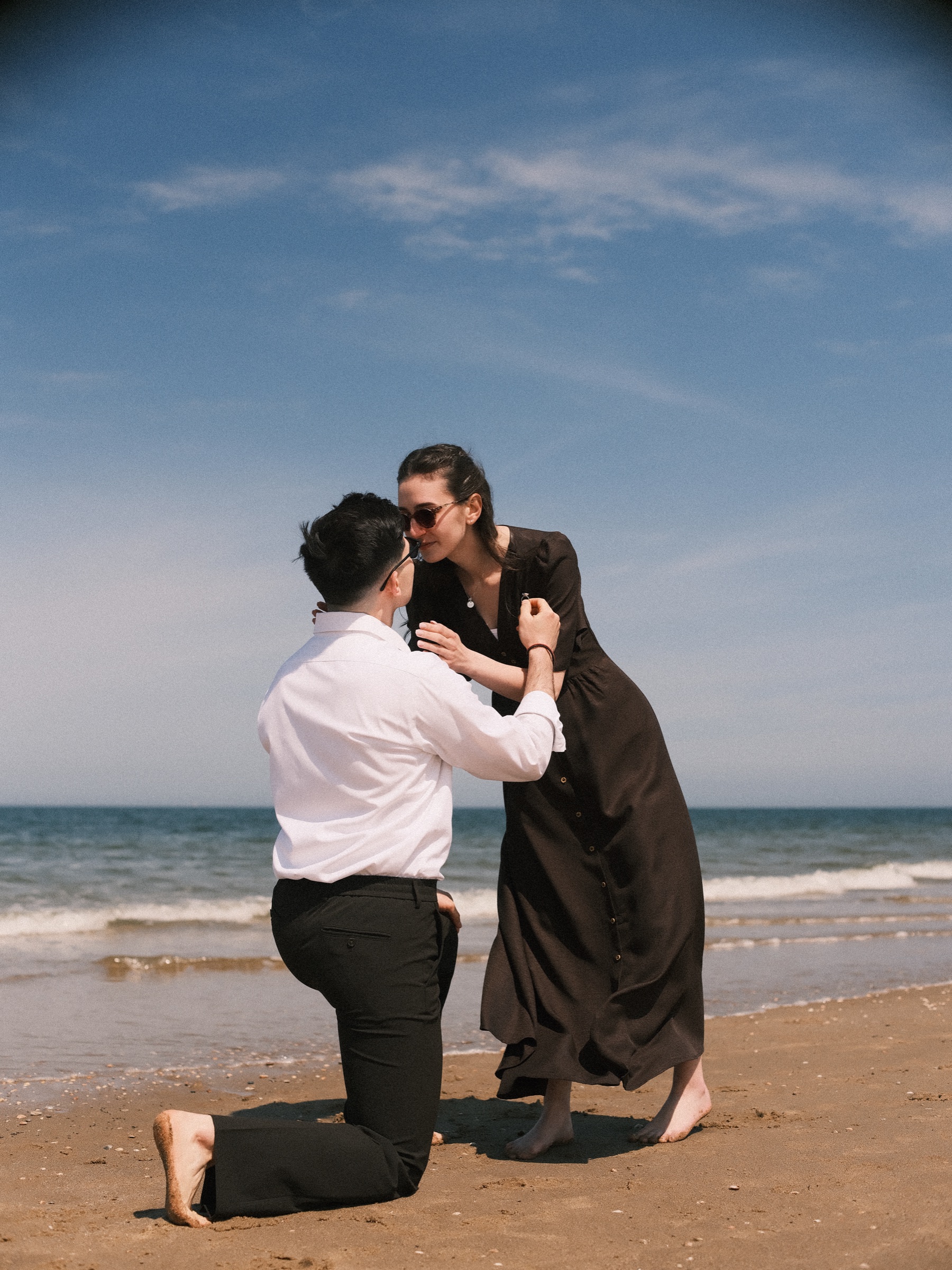 Woman in sunglasses bending down to kiss the man still on one knee in the sand, blue sky and small waves behind them