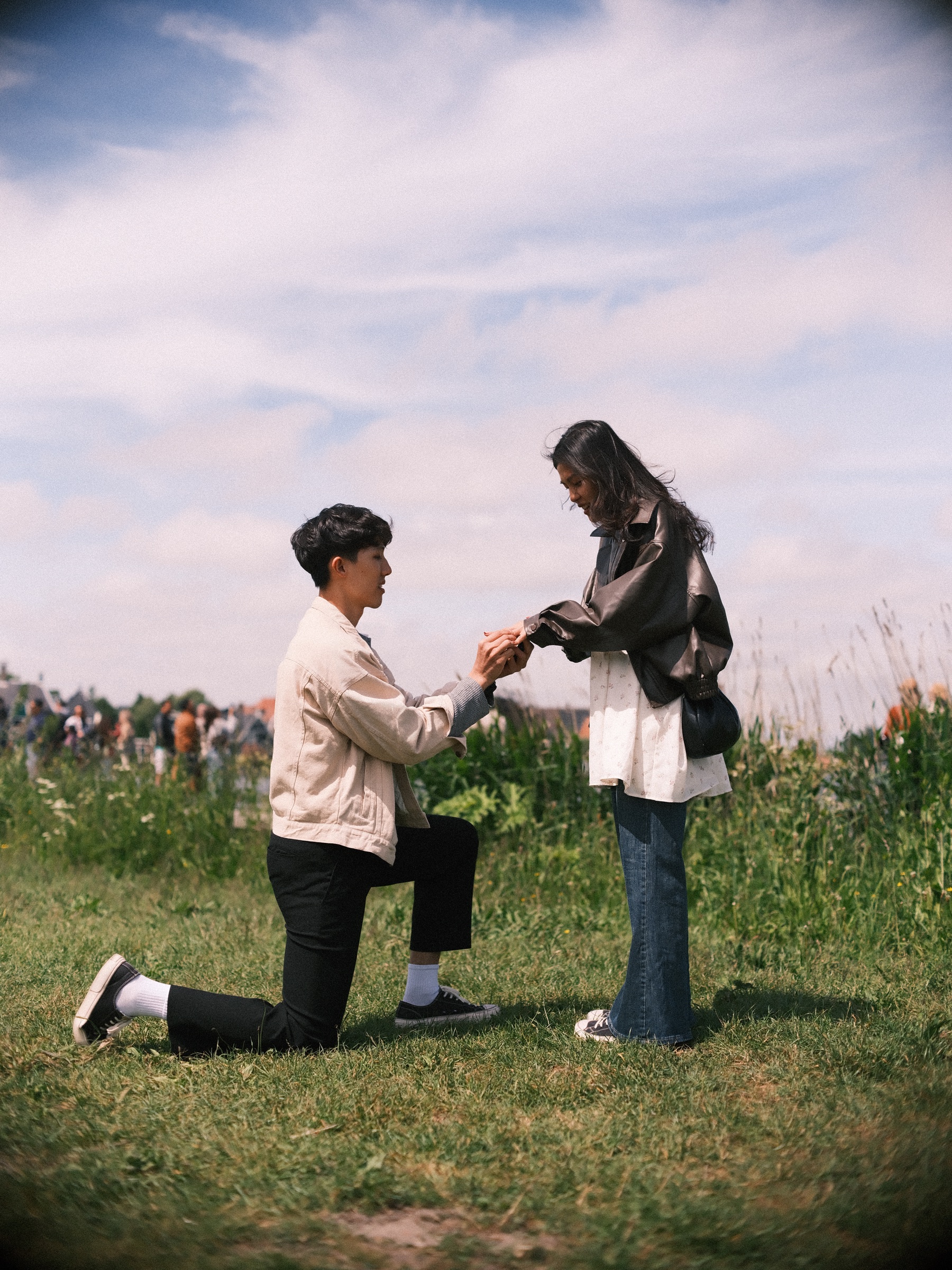 Kneeling man holding her hand with the ring as she looks down, wind in her long hair and visitors far behind under a wide sky