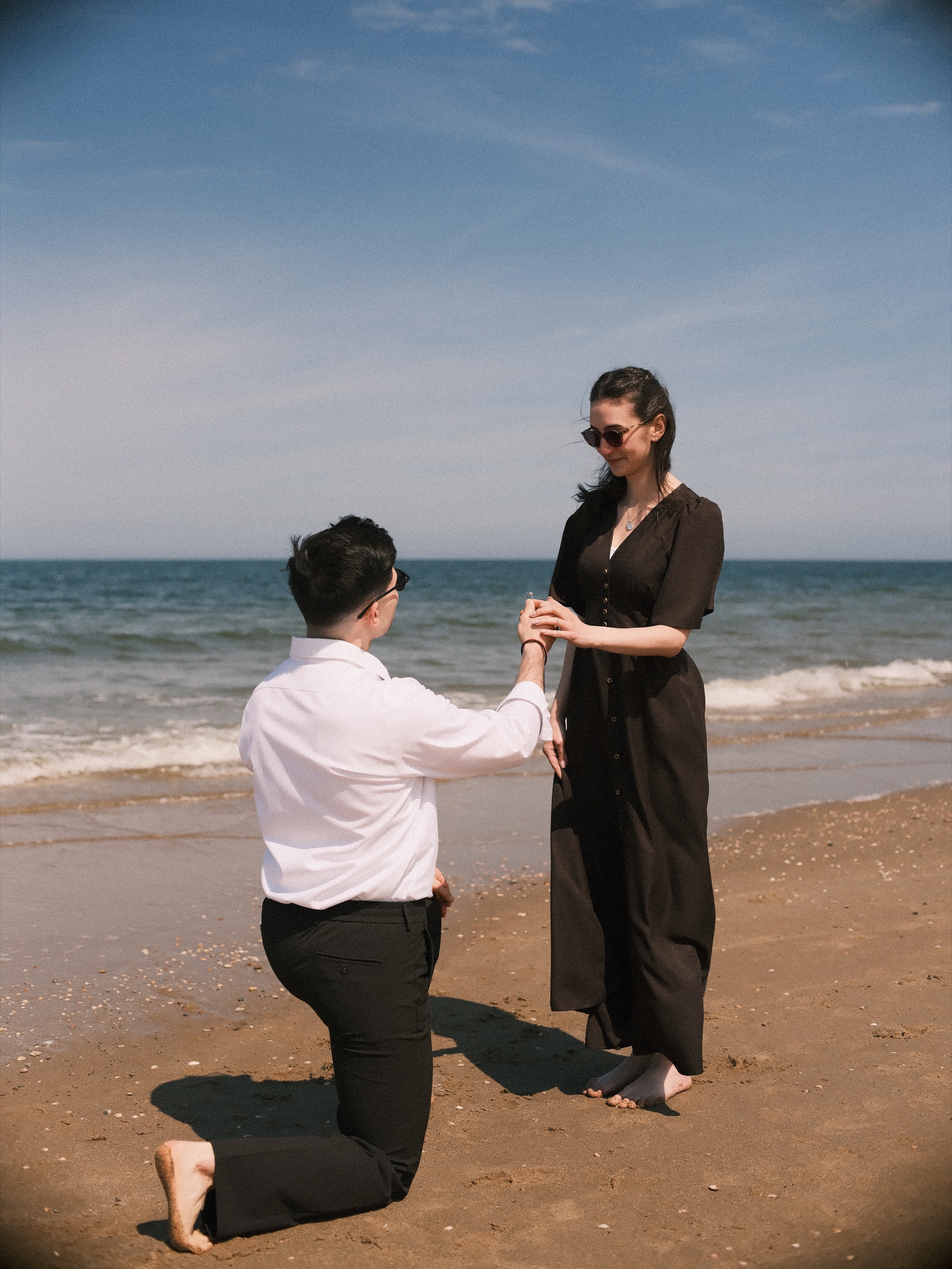 Kneeling man holding his partner's hand at the waterline, the ring ready, her long dark dress moving in the sea breeze