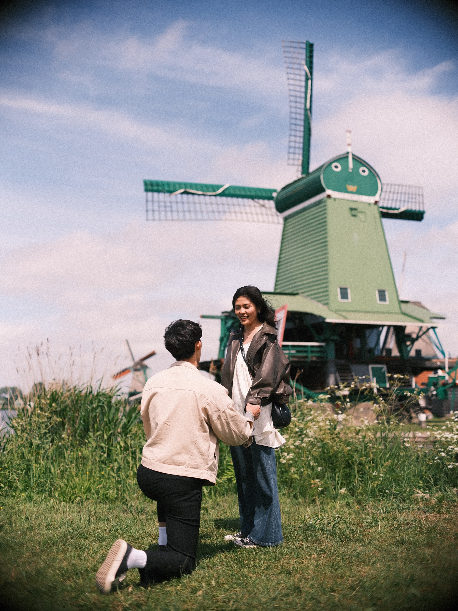Man in a cream jacket kneeling on the grass before a green windmill as she smiles down at him, a proposal at the Zaanse Schans