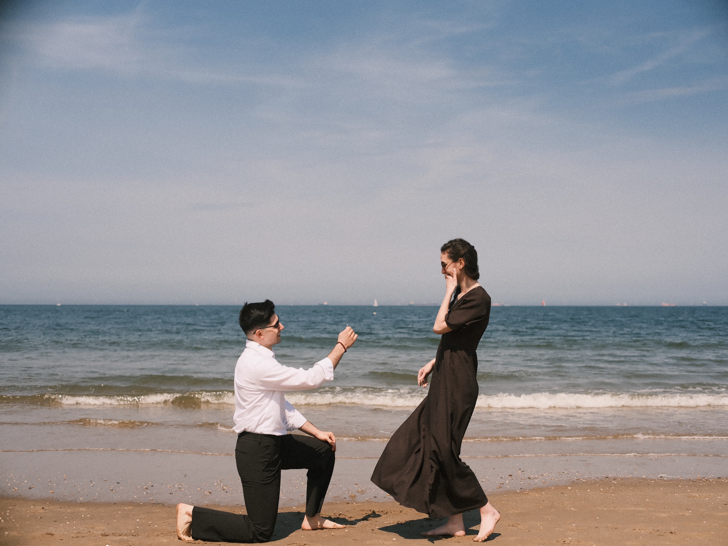 Man in a white shirt kneeling in the sand as she lifts a hand to her face in a long brown dress, a proposal on Scheveningen beach