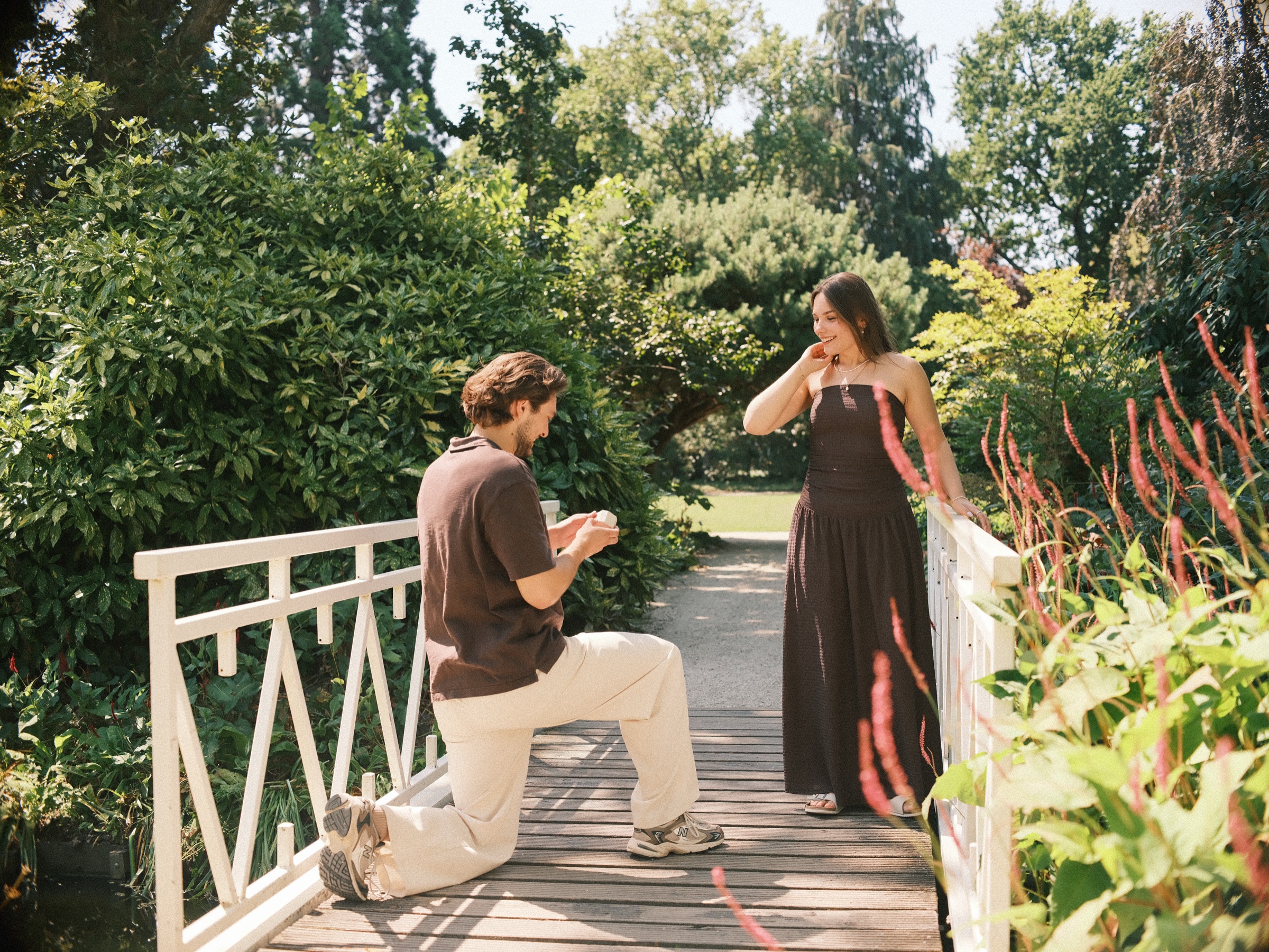 He kneels and holds up the ring on the white bridge as she lifts a hand to her mouth, red flowers in front, a surprise proposal in Rotterdam