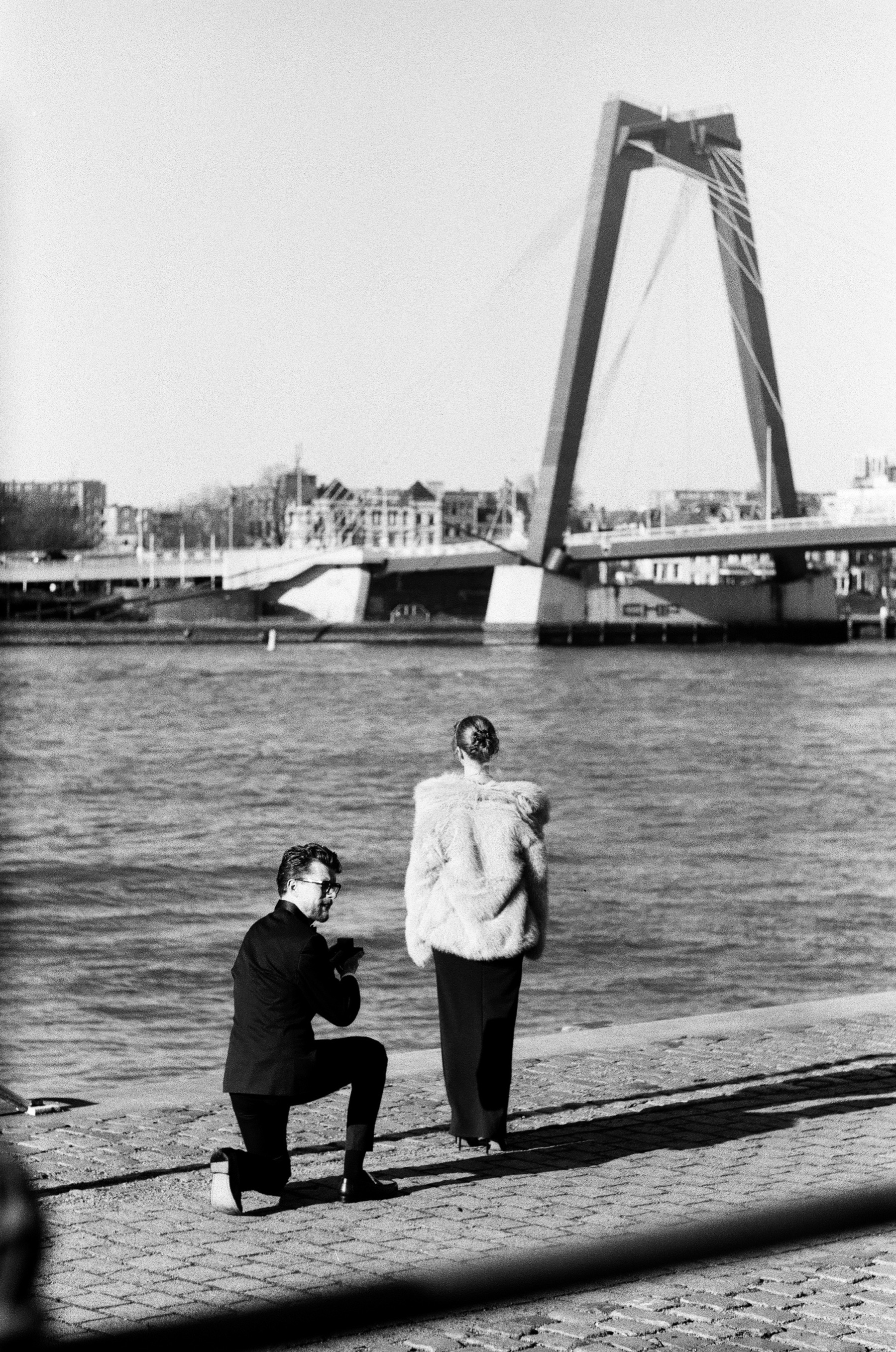 Man in a tuxedo kneeling on the cobbled quay as she stands by the Maas, the Willemsbrug behind them, a proposal on black and white film