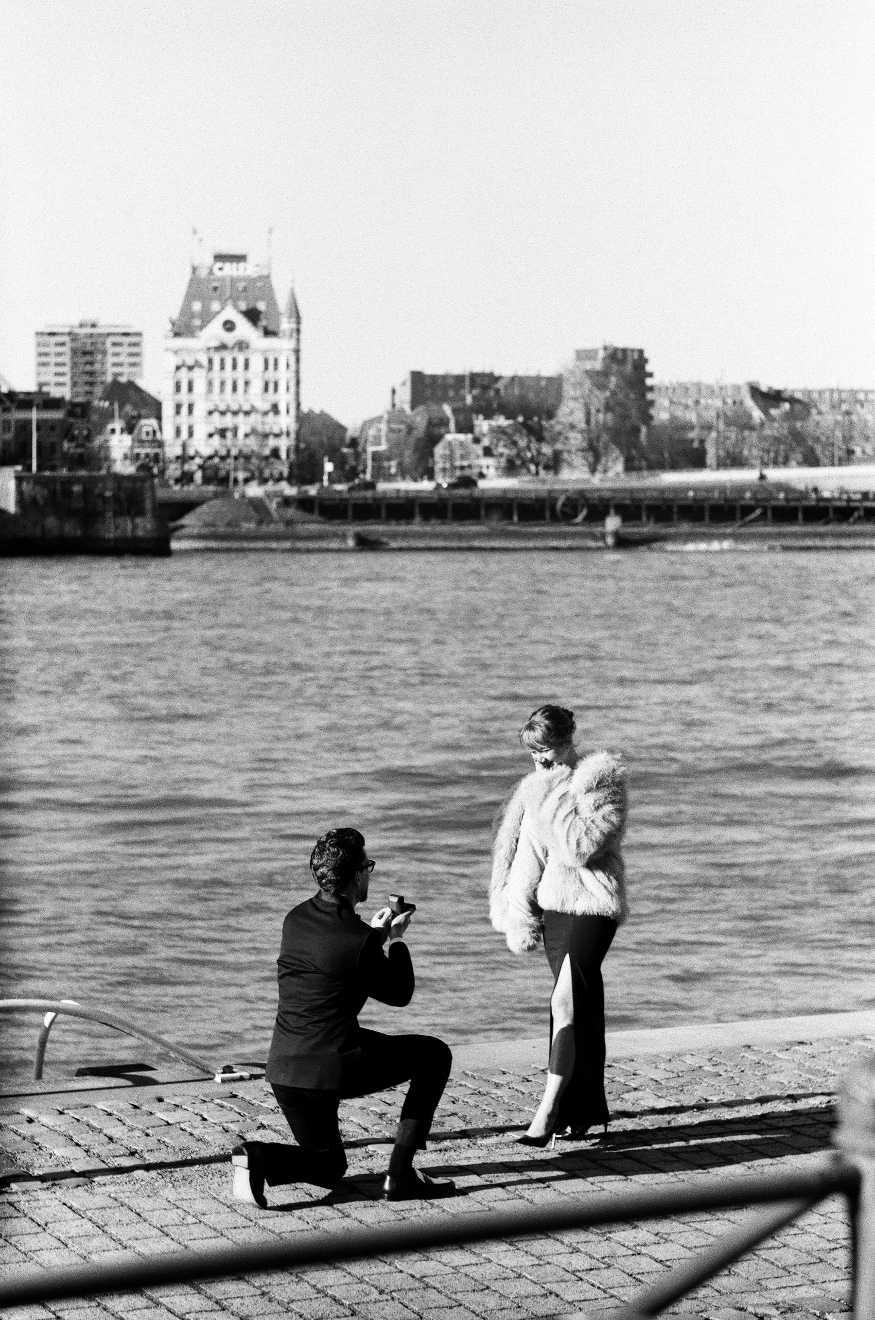 From across the cobbled quay, he kneels before her by the Maas with Het Witte Huis on the far bank, a surprise proposal on black and white film