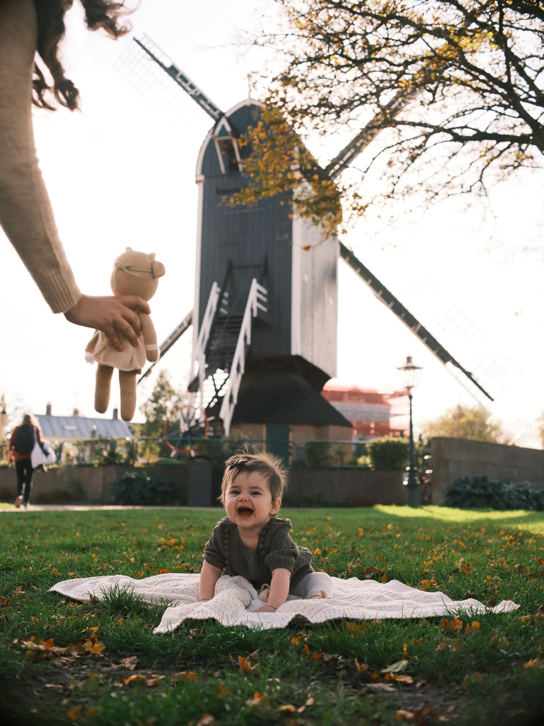 A hand dangles a knitted toy above the lens while the baby on the blanket laughs up at it, the windmill and autumn trees behind
