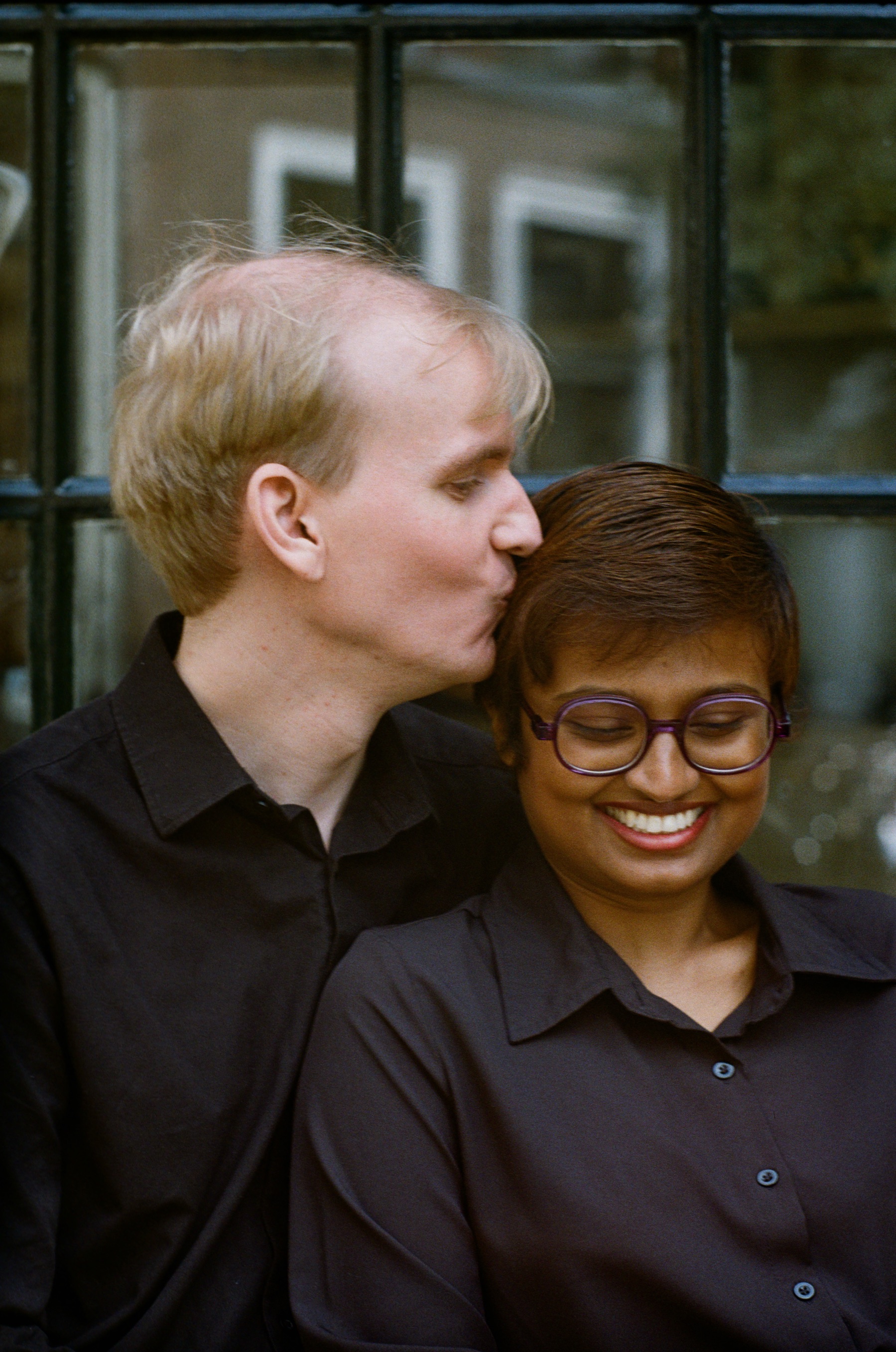 He kisses her temple as she smiles widely behind round glasses, both in black shirts, a close frame on Kodak Gold in Leiden