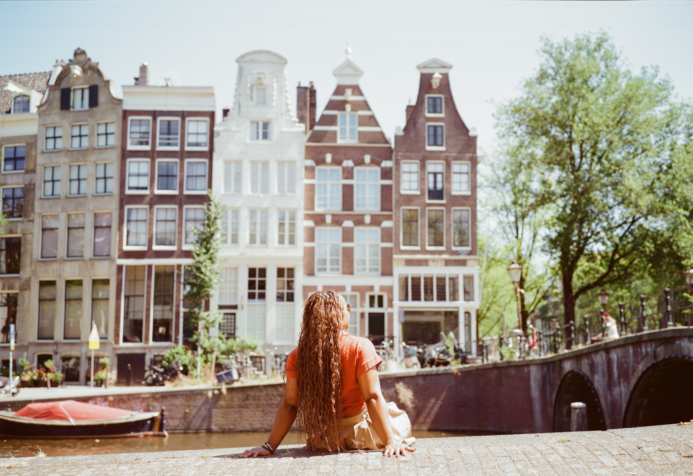 Woman with long copper-brown braids sitting on a canal quay in Amsterdam, her back to the camera, facing gabled houses and a stone bridge