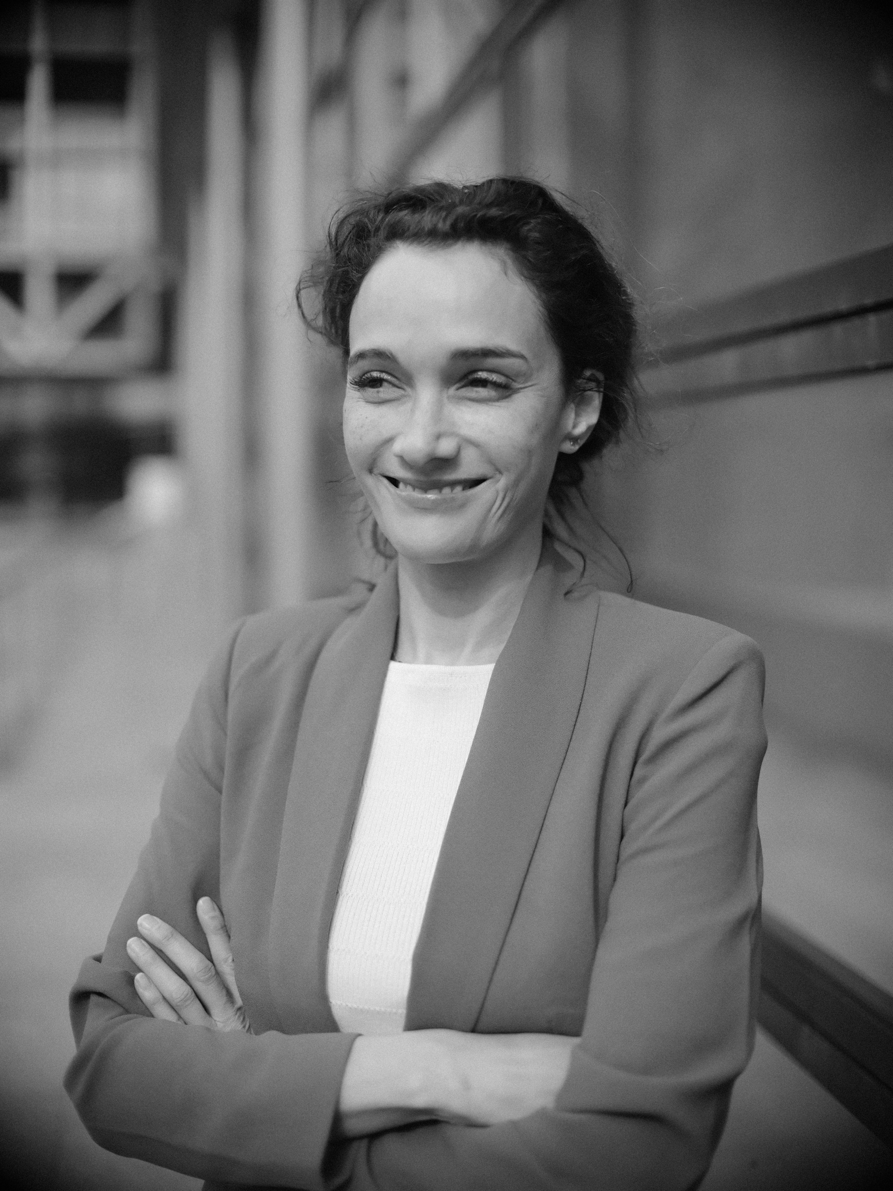 Zwart-witportret van een lachende advocaat in een colbert en wit topje, armen over elkaar, met een modern gebouw zacht erachter