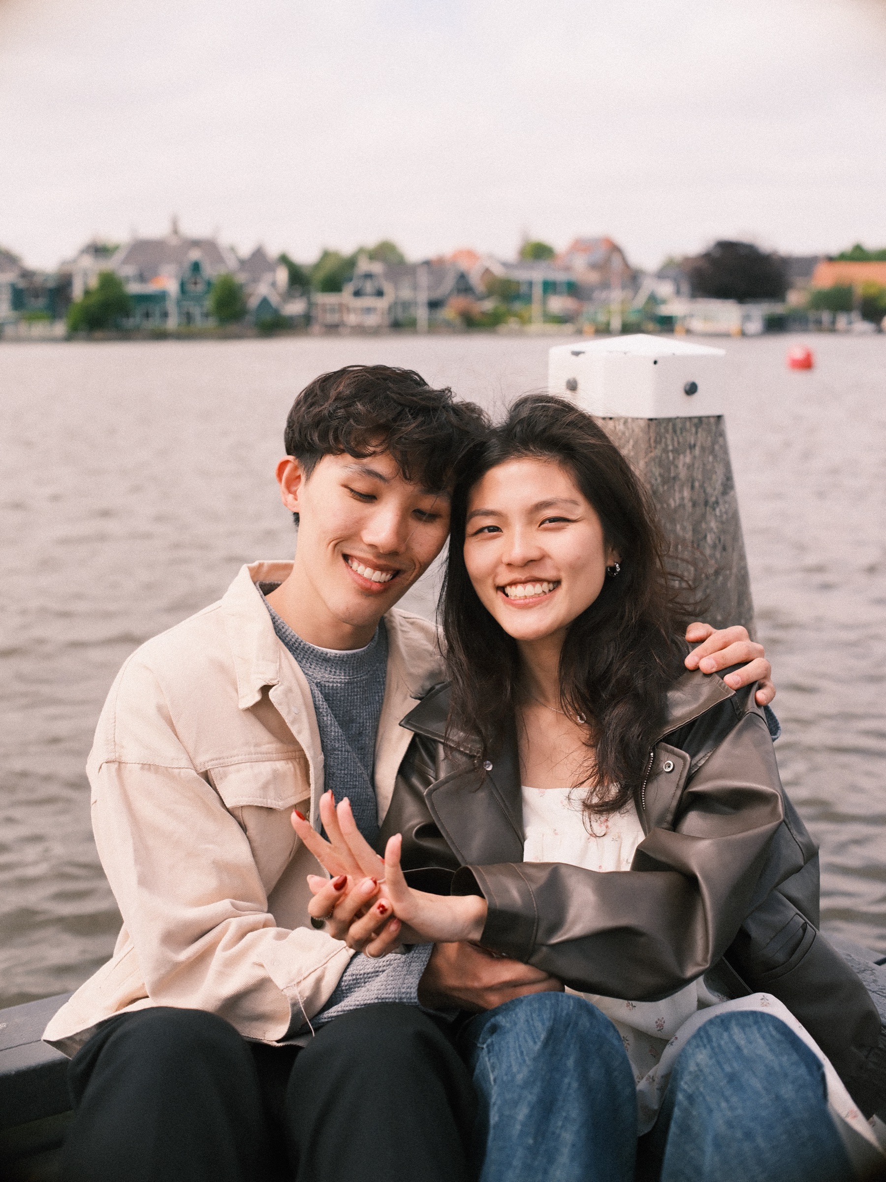 Newly engaged couple sitting cheek to cheek at the end of the jetty and laughing, her hand on his, green houses across the river