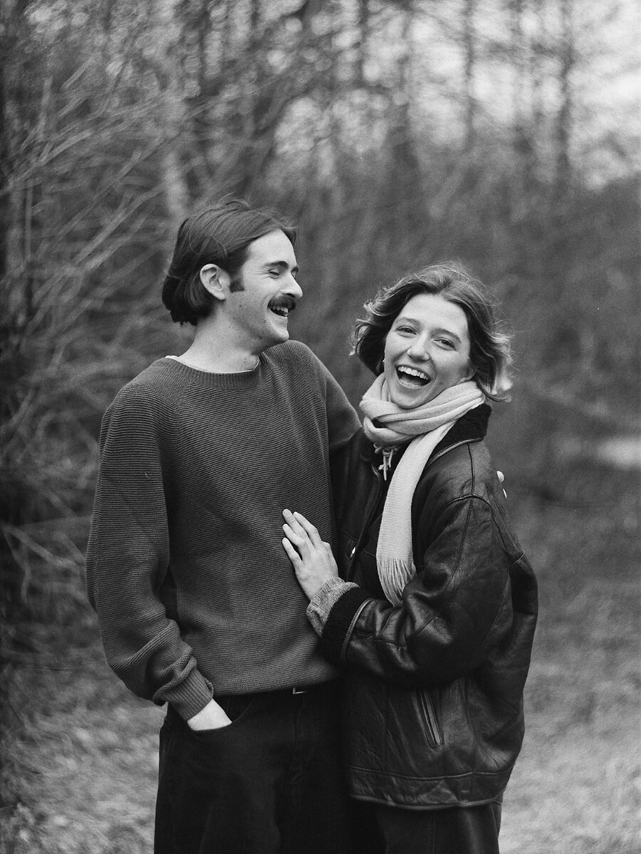 Laughing together among the trees, a couple stand close with her hand on his jumper in a black-and-white film portrait