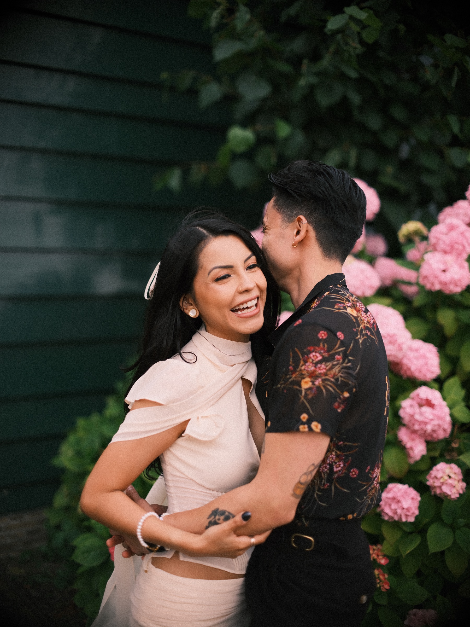Woman in an ivory top with a long ribbon laughing in her partner's arms in front of pink hydrangeas and a dark green wooden wall