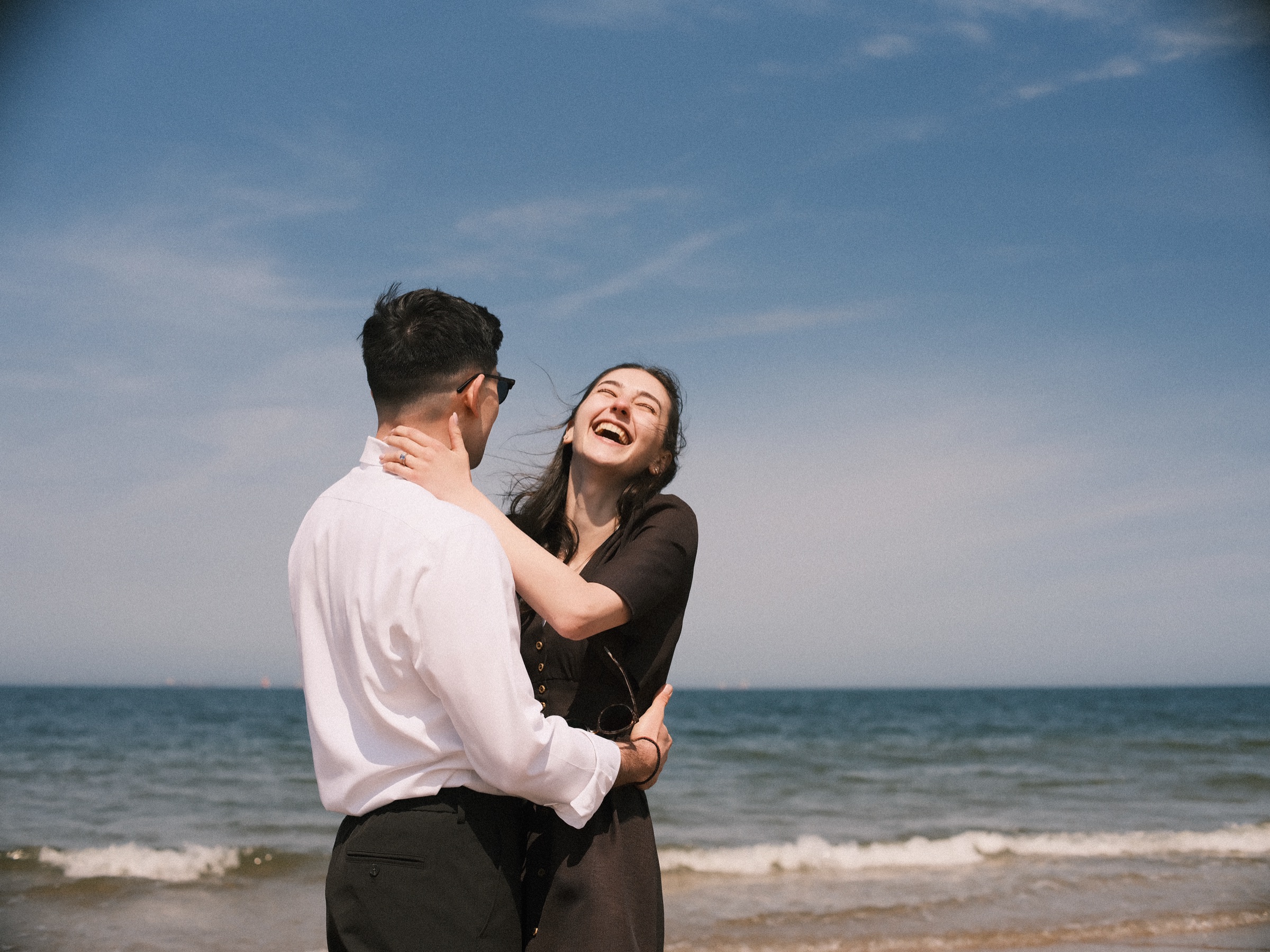 Couple hugging and laughing by the sea right after the proposal on Scheveningen beach, her hand on the back of his neck