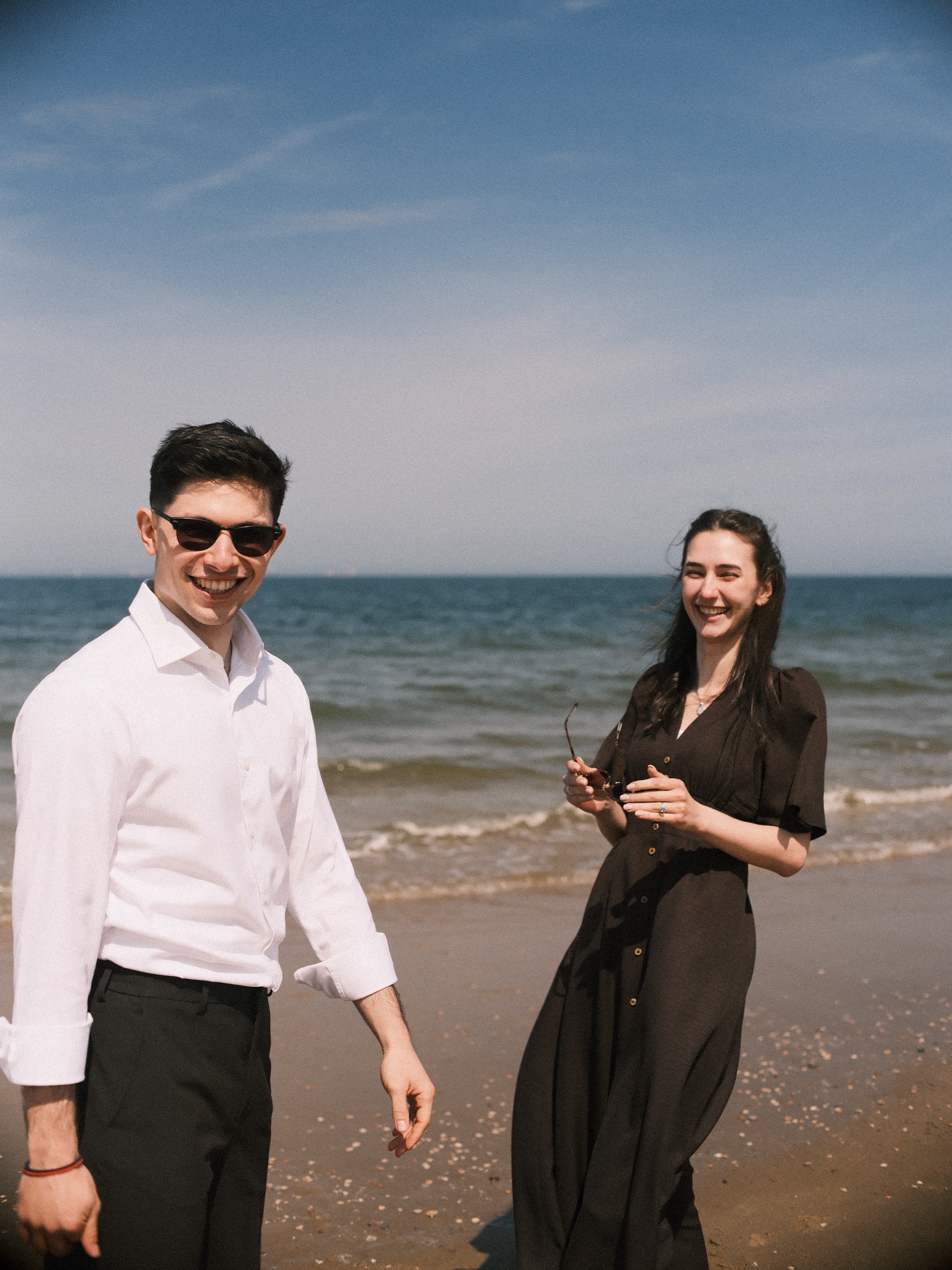 Grinning in sunglasses and a white shirt, he walks toward the lens while she laughs beside him on the beach, the sea bright behind them