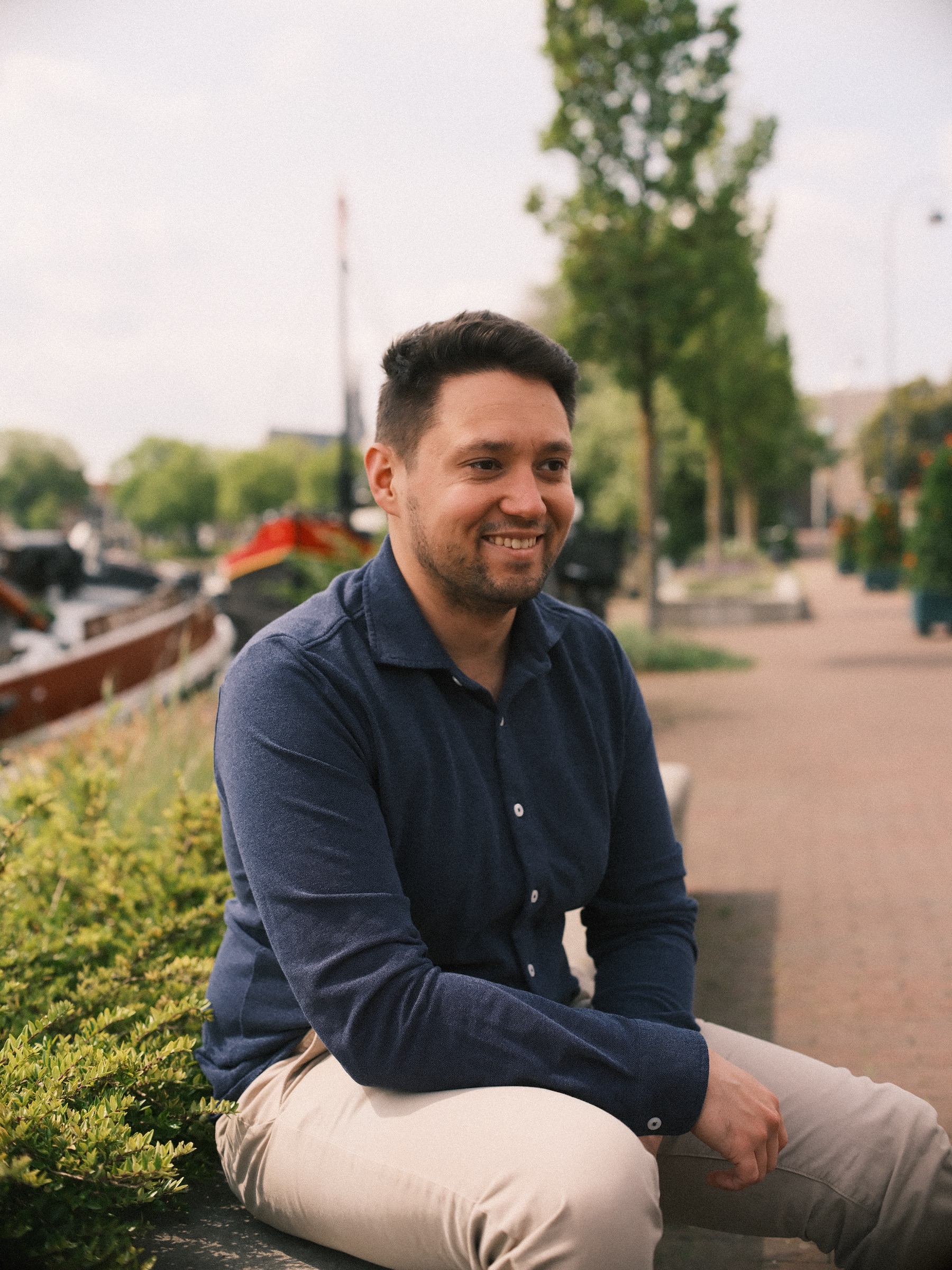 Lachende man in donkerblauw overhemd zit aan het water en kijkt opzij, naast groene struiken en een aangemeerde boot