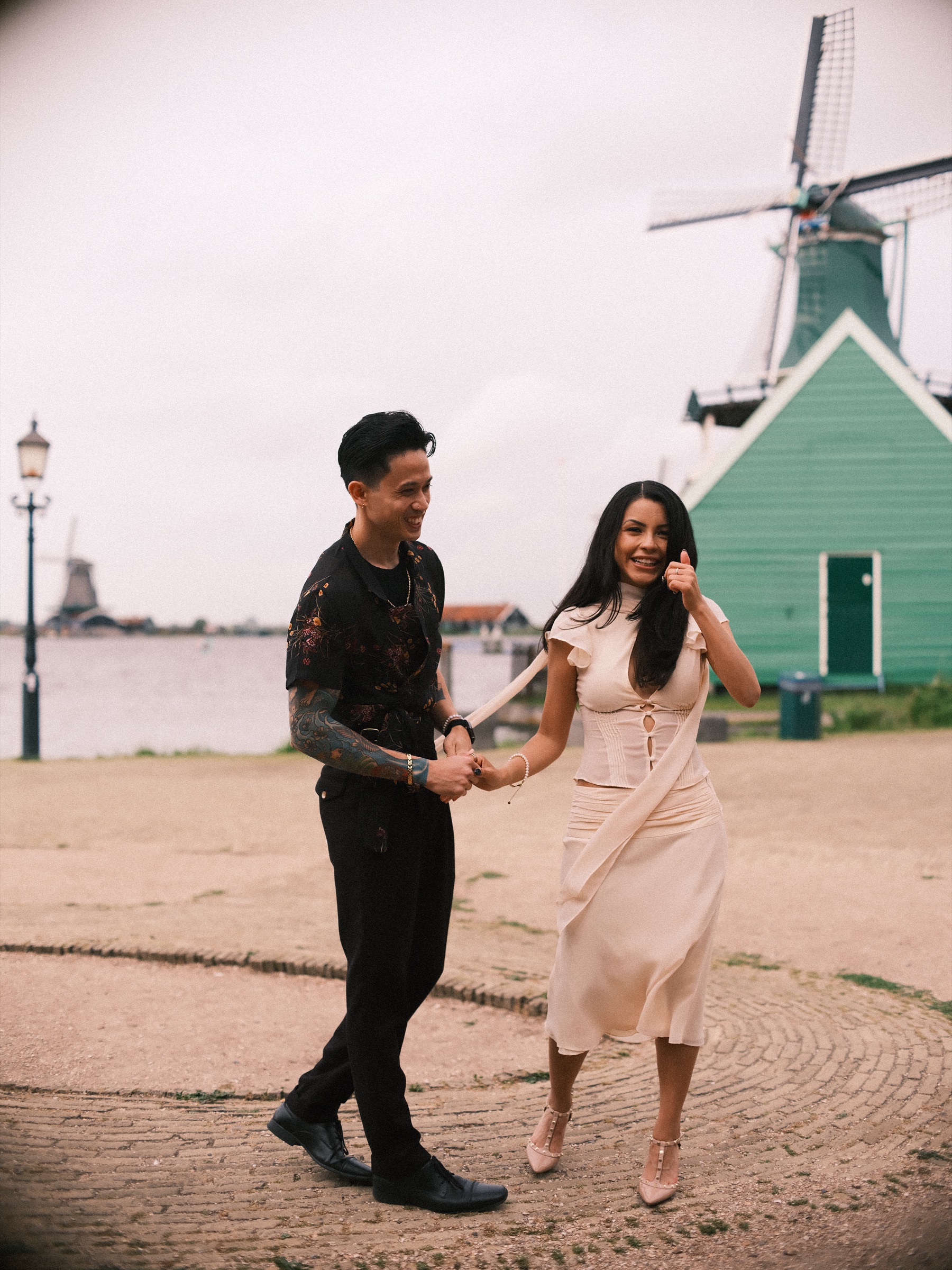 Laughing woman in an ivory skirt and nude heels stepping toward her partner as he leads her by the hand, the river and a lamppost behind