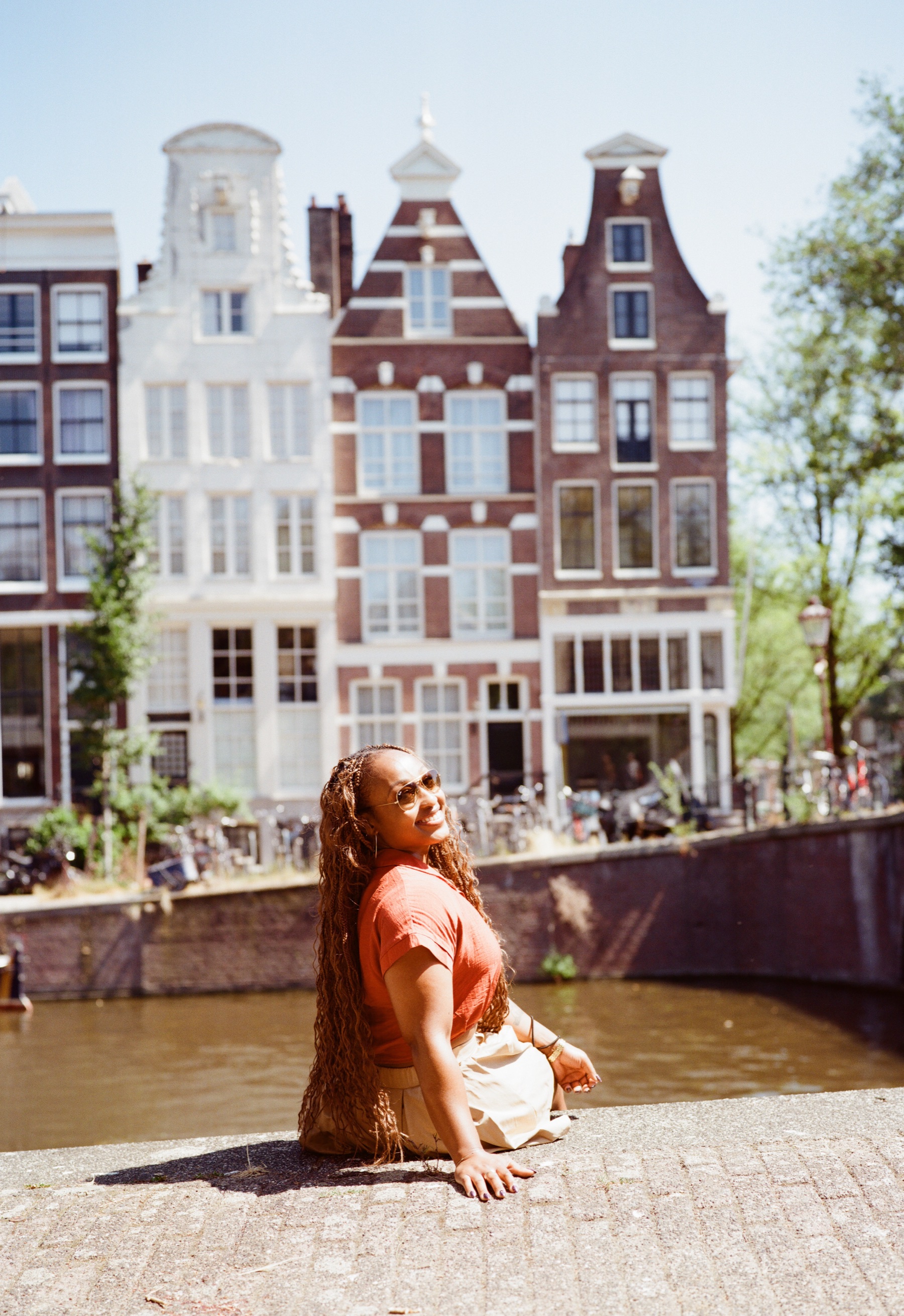 Lachende vrouw in een oranje blouse met zonnebril zit op de kade en kijkt over haar schouder, met grachtenpanden achter haar