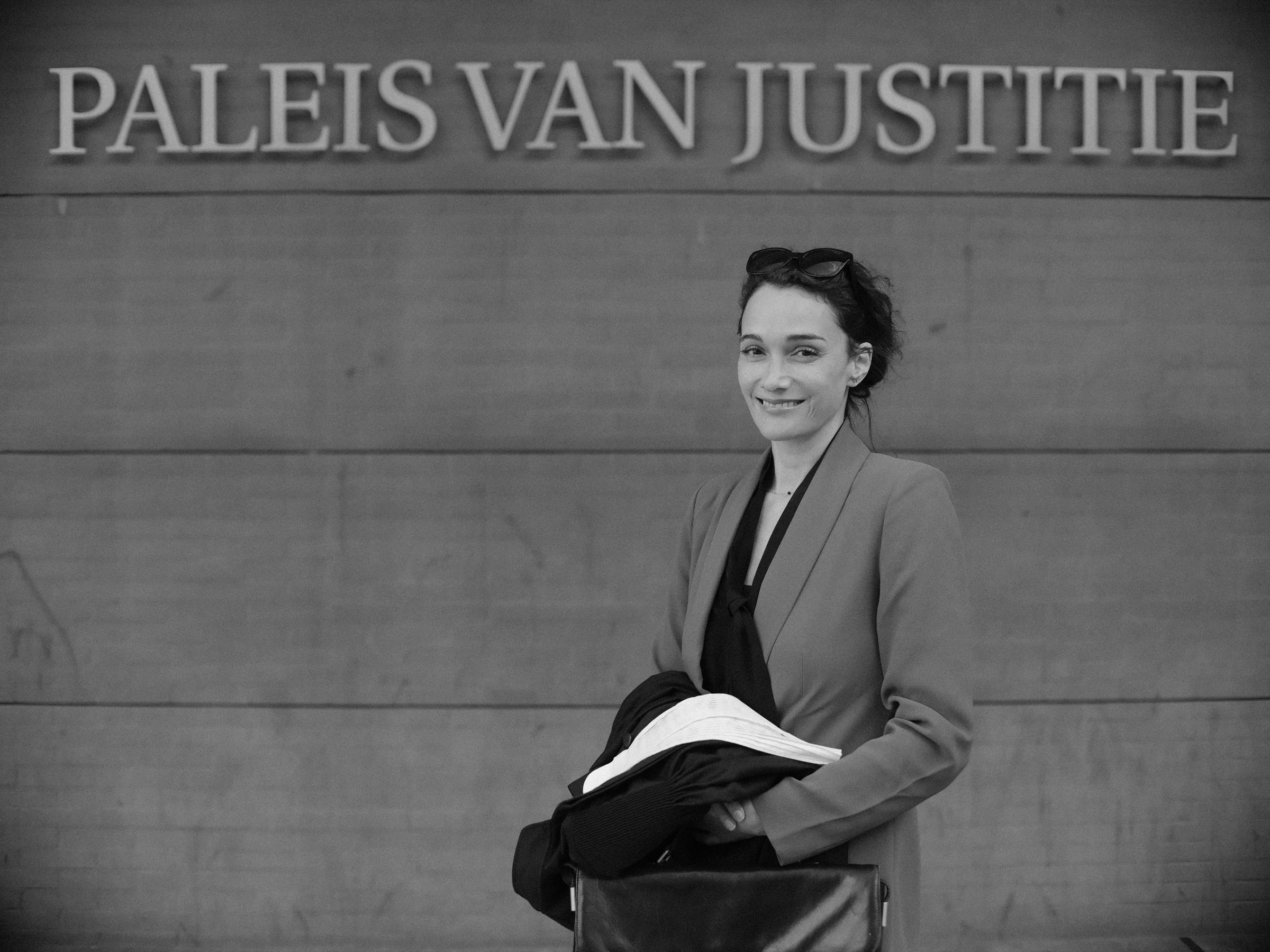Advocaat lacht onder de letters van het Paleis van Justitie in Den Haag, met haar toga en witte bef over de arm, in zwart-wit