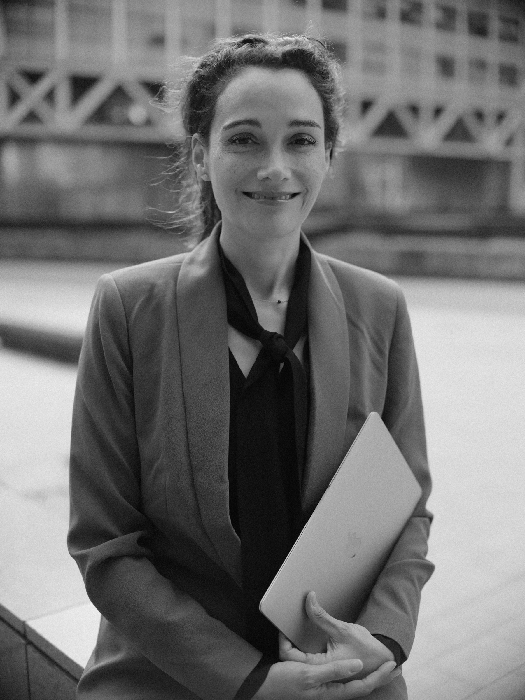 Advocaat met een laptop in haar handen, in een colbert en zwarte strikblouse, glimlacht in zacht zwart-wit voor een modern gebouw