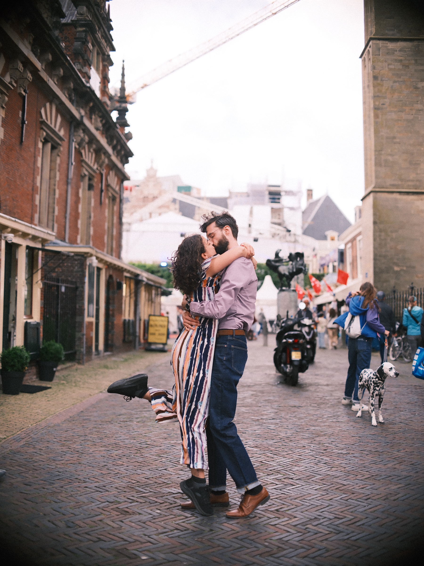 Stel kust terwijl zij een voet omhoog schopt op de klinkers bij de Grote Markt in Haarlem, met een dalmatiër die op een zomerdag voorbijloopt